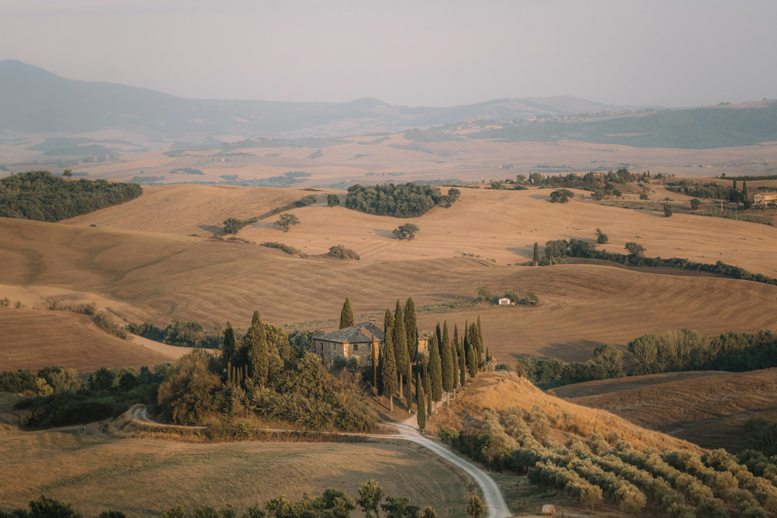 Rolling hills and countryside landscape in Tuscany, Italy with sparse trees, a winding dirt road, and a small stone farmhouse surrounded by cypress trees.