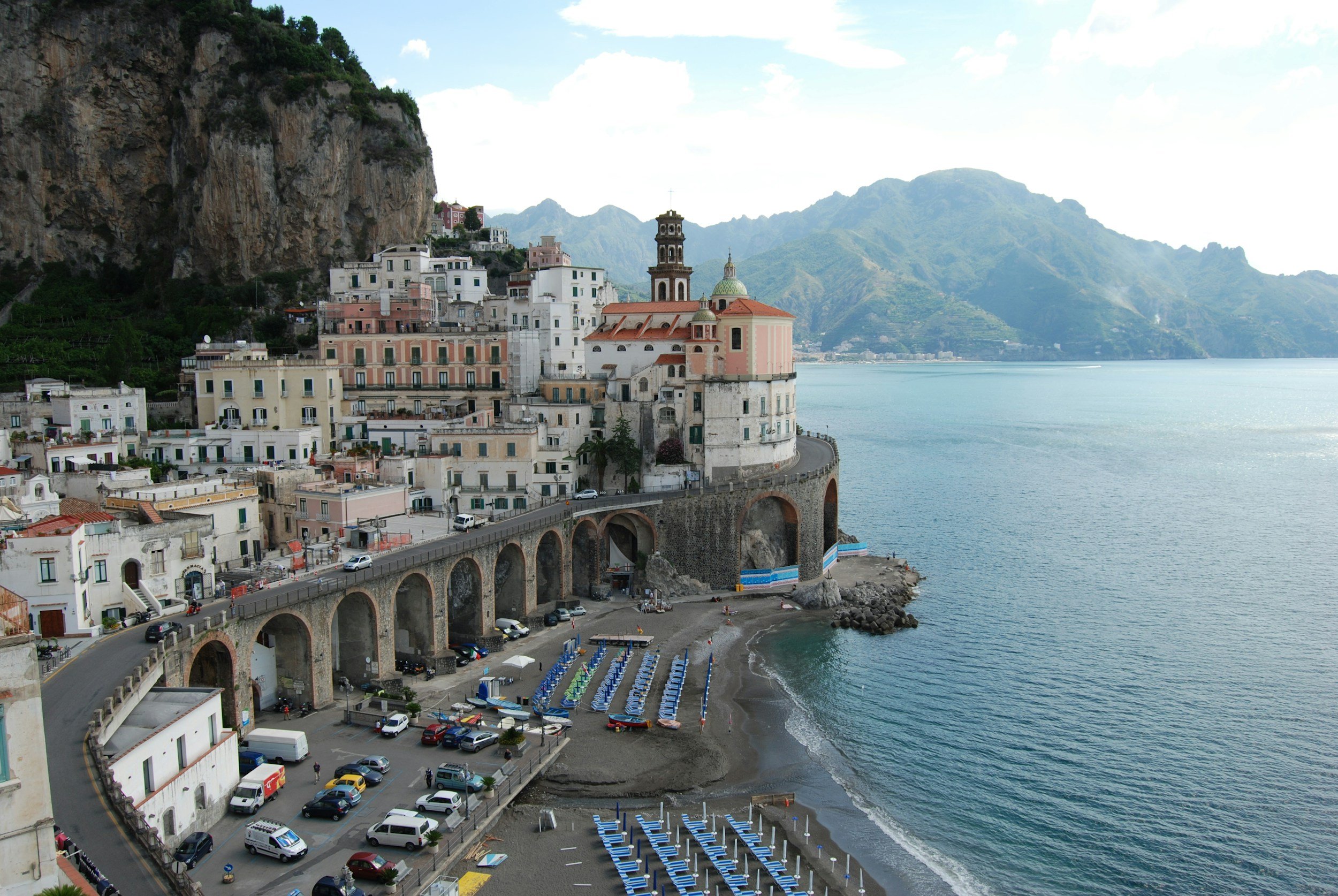 Coastal town with colorful buildings, a church with a bell tower, and a seaside beach with umbrellas and chairs, overlooking a bay with mountains in the background.