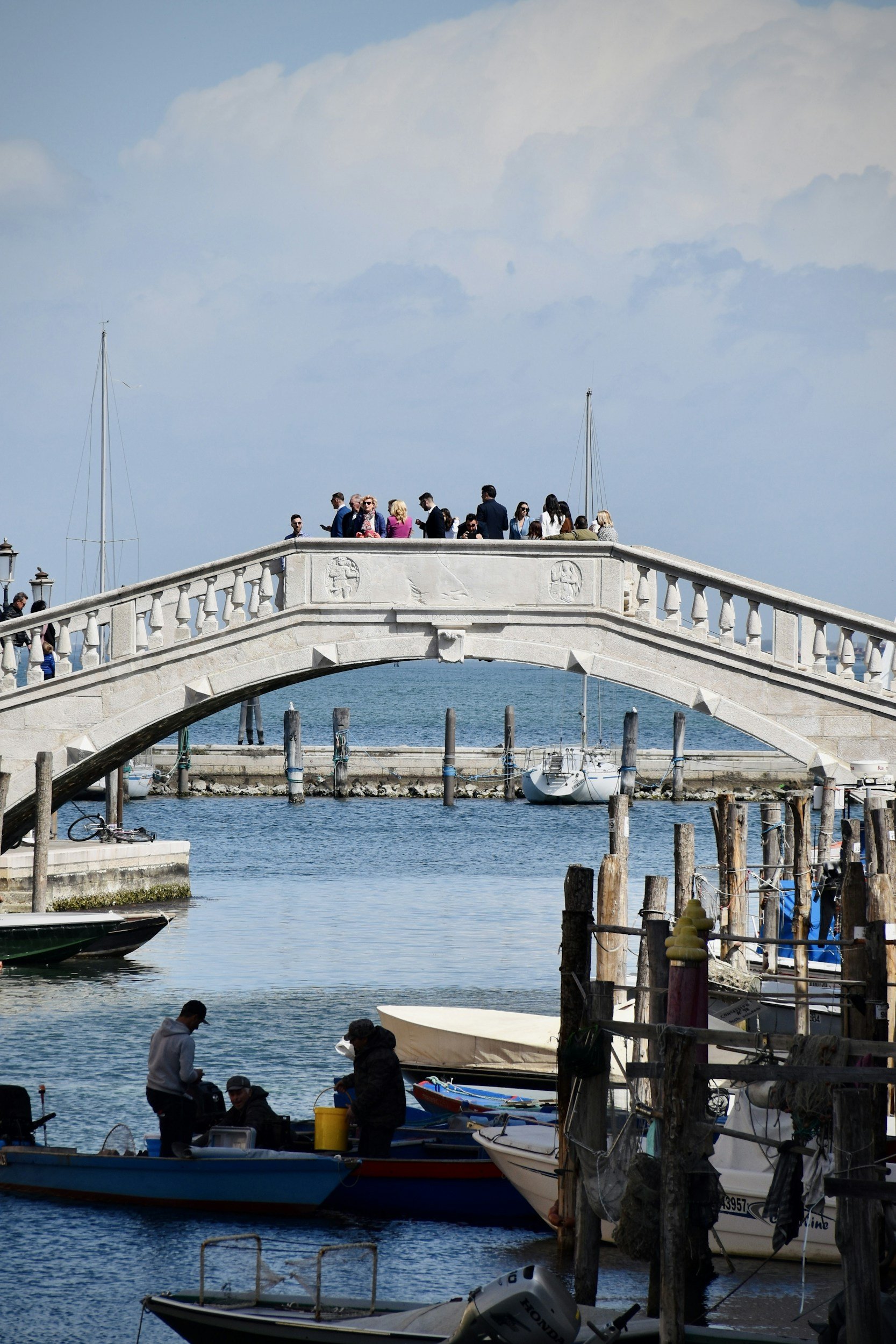 People gathering on a stone bridge over water with boats docked below, sailboats in the background, and a partly cloudy sky.