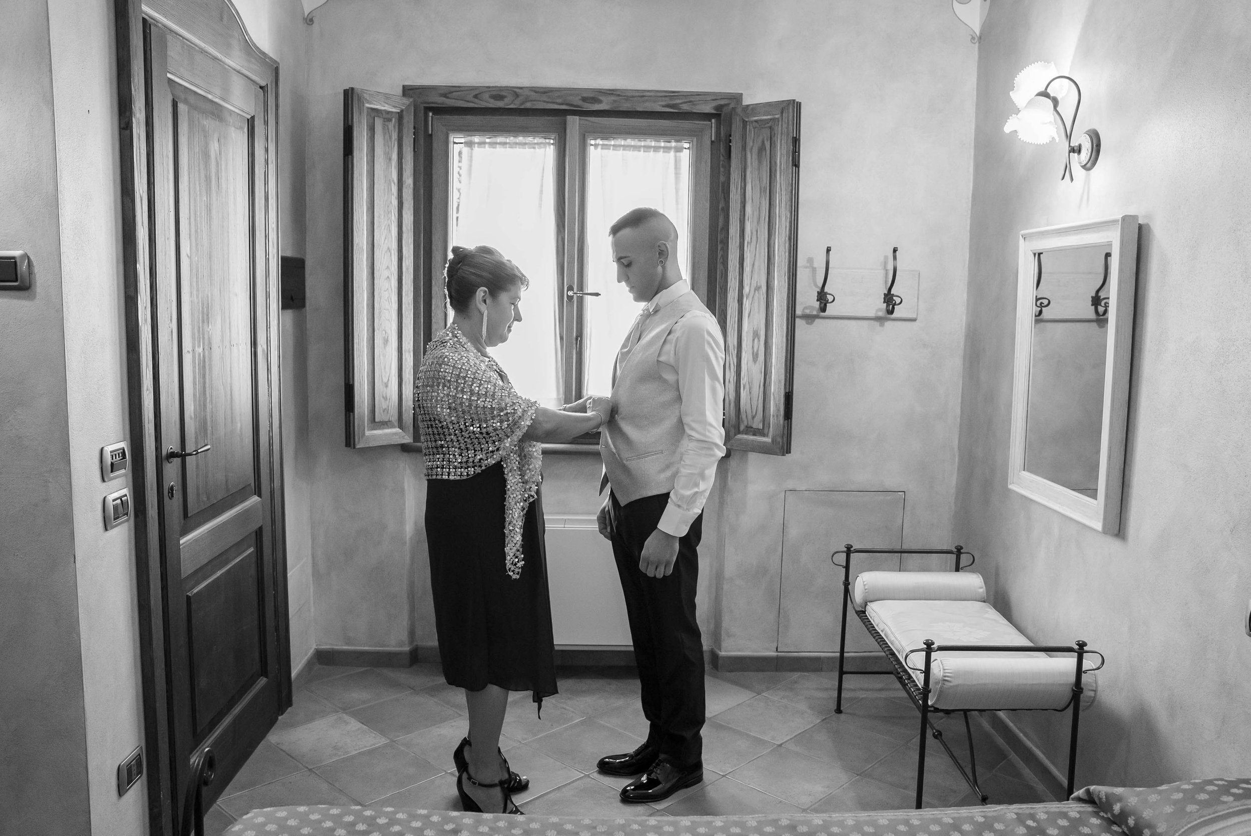 A woman and a man are standing in a room near a window. The woman appears to be adjusting or fastening the man's jacket. The room has a wall-mounted lamp, a mirror on the wall, and a bench with a cushion. The scene is in black and white.