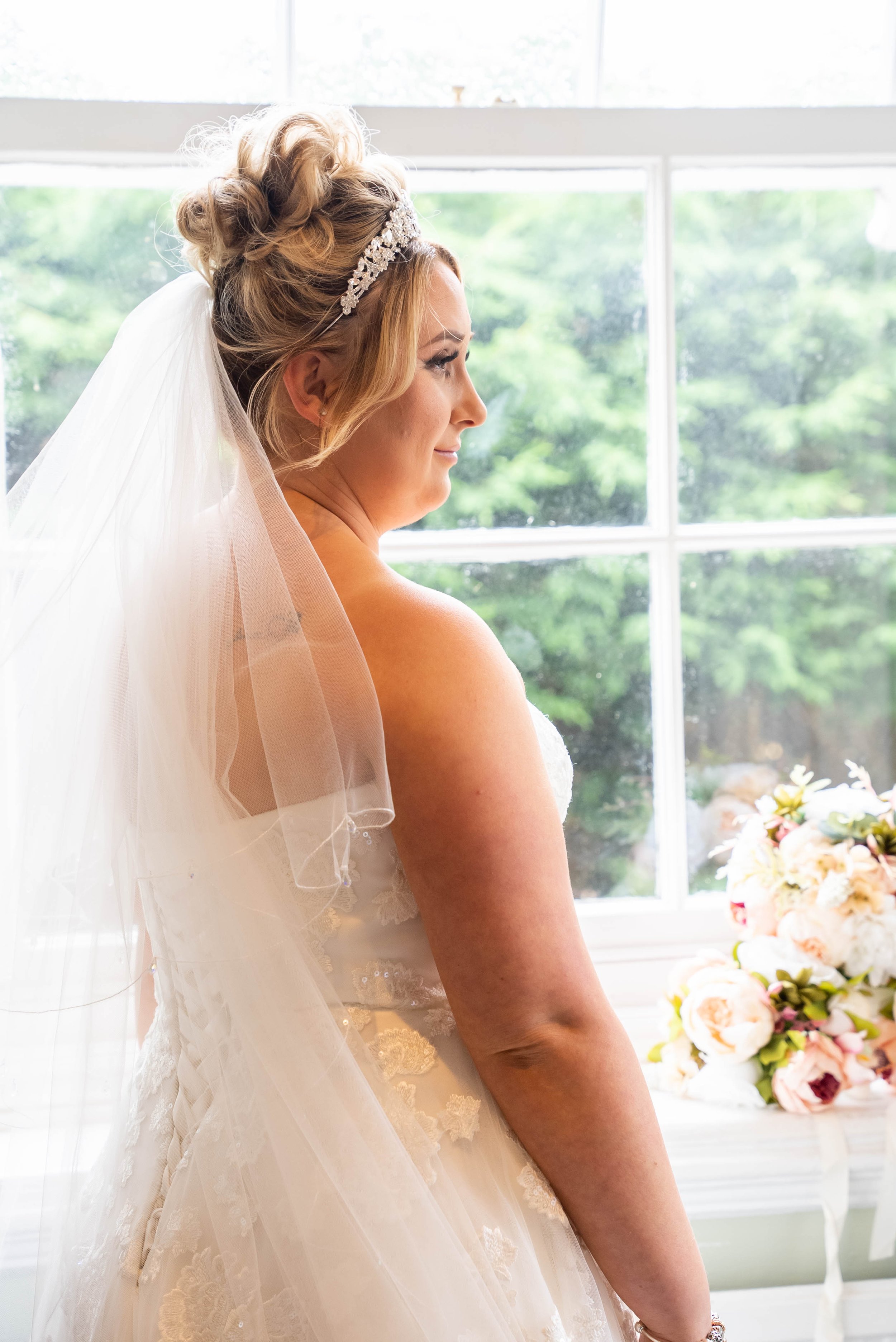 A bride stands indoors near a large window with green trees outside, wearing a wedding dress, veil, and tiara, with a bouquet of flowers on a table nearby.