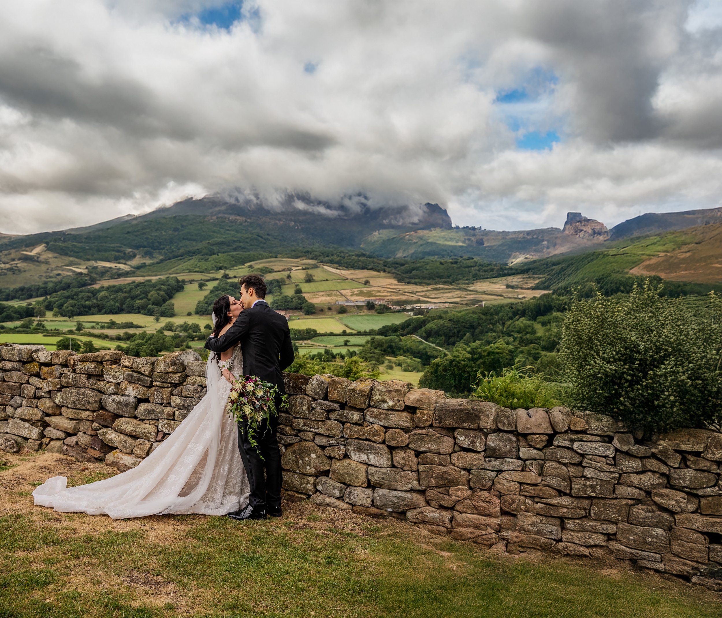 A bride and groom embrace outdoors in front of a stone wall, with green hills and mountains under a cloudy sky in the background.