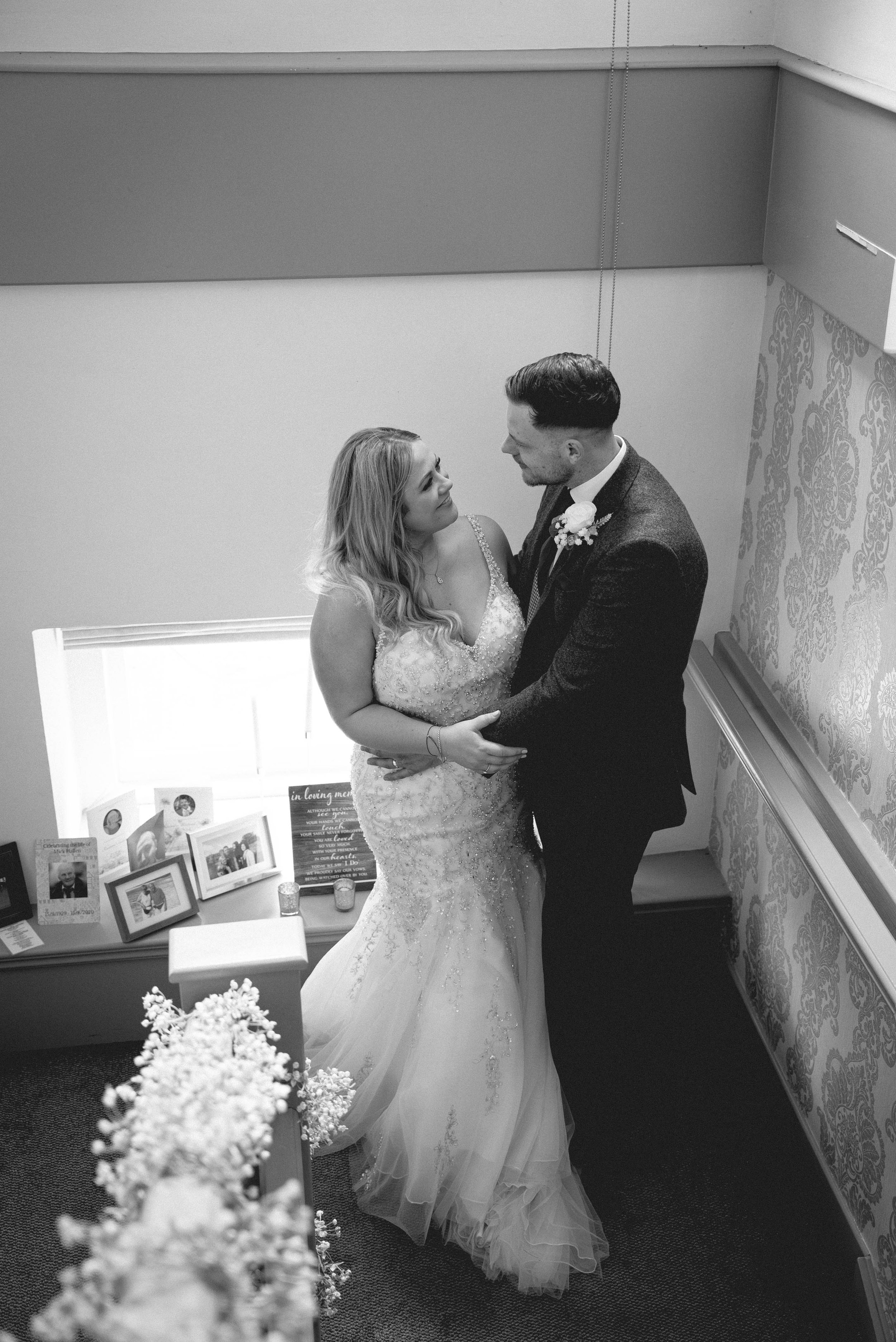 A bride and groom embracing on their wedding day, standing indoors near a decorated wall and table with framed photos and flowers.