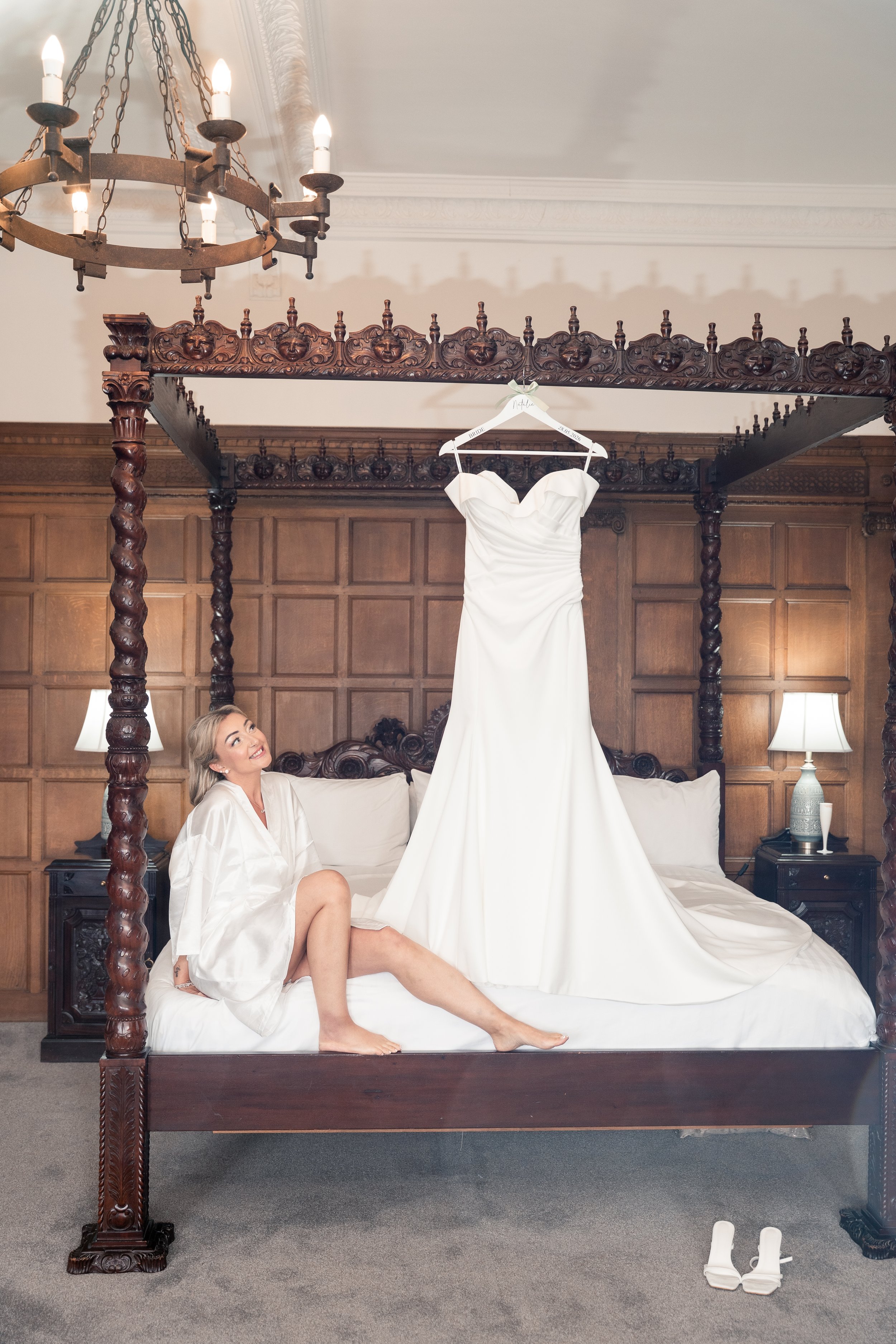 A woman with blonde hair is sitting on a bed, wearing a white satin robe, smiling and looking up. A white wedding dress is hanging from a carved wooden canopy bed behind her, and a pair of white high-heeled shoes are on the floor in front of the bed.