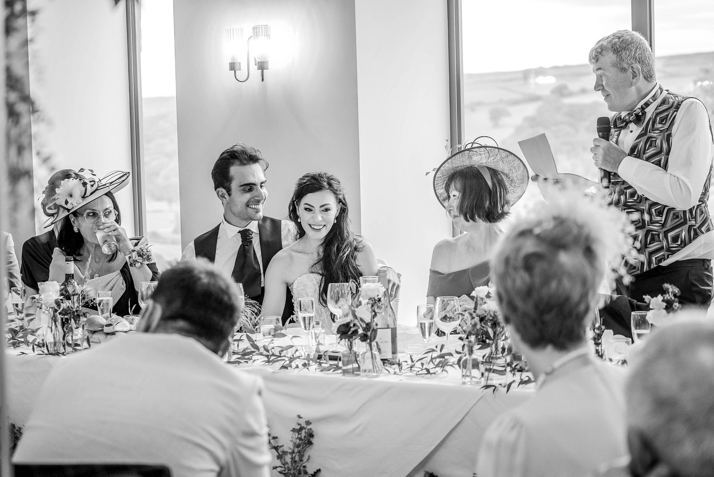 A wedding reception with a bride and groom sitting at a decorated table, listening to a man giving a speech with a microphone. The bride has long hair and the groom has dark hair, both are smiling. Several guests, mostly women, are seated around the 