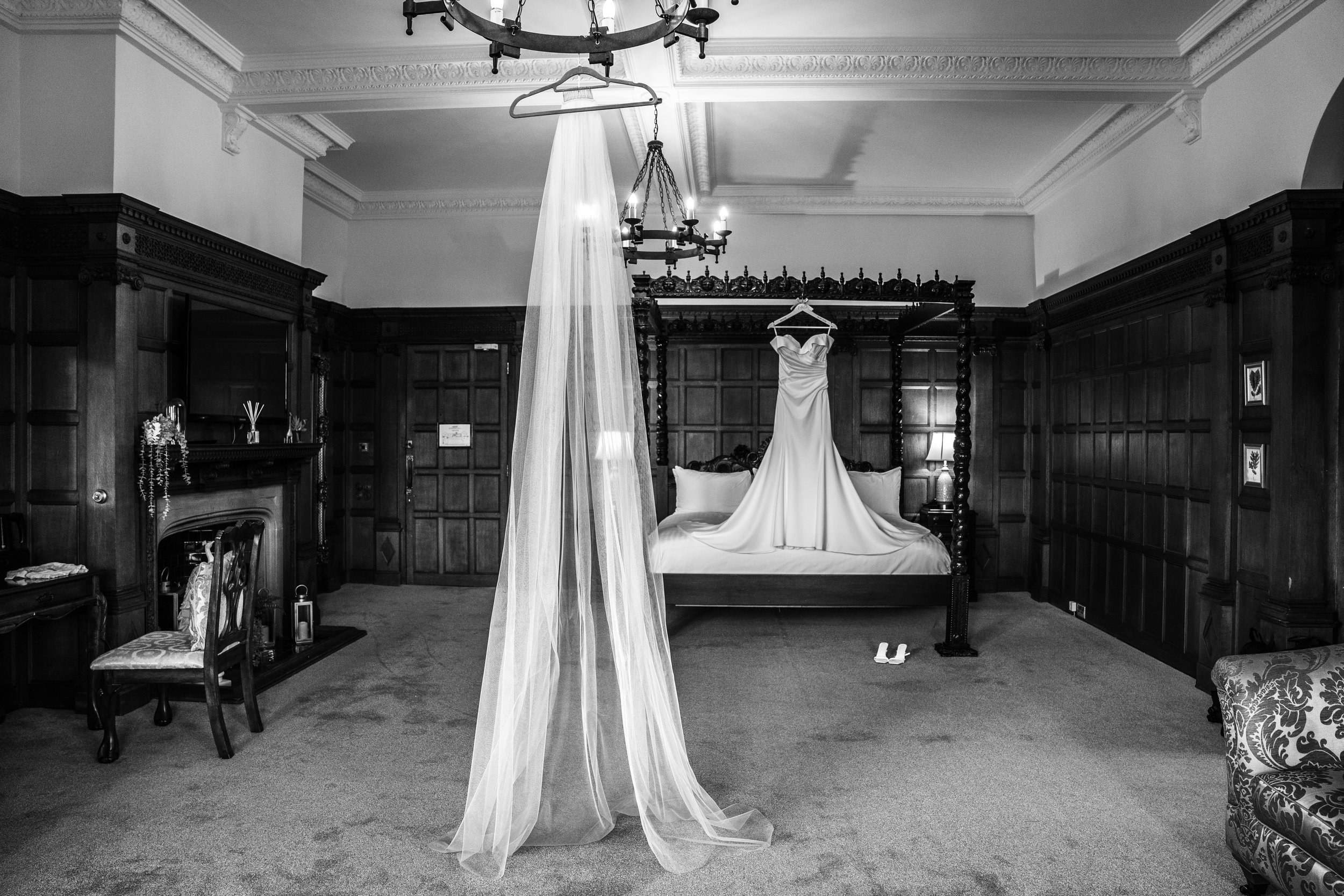 A wedding dress hanging from a four-poster bed, in a room with wood paneling and ornate crown molding. A sheer curtain is draped from the ceiling, partially covering the dress, and a chandelier hangs overhead.