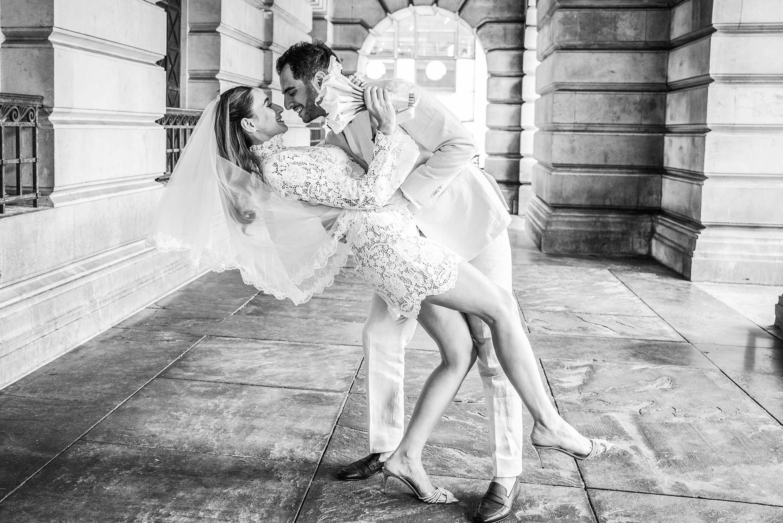 A bride and groom dancing affectionately inside a grand stone building, with the groom lifting the bride as she leans back in a joyful pose, both smiling at each other.