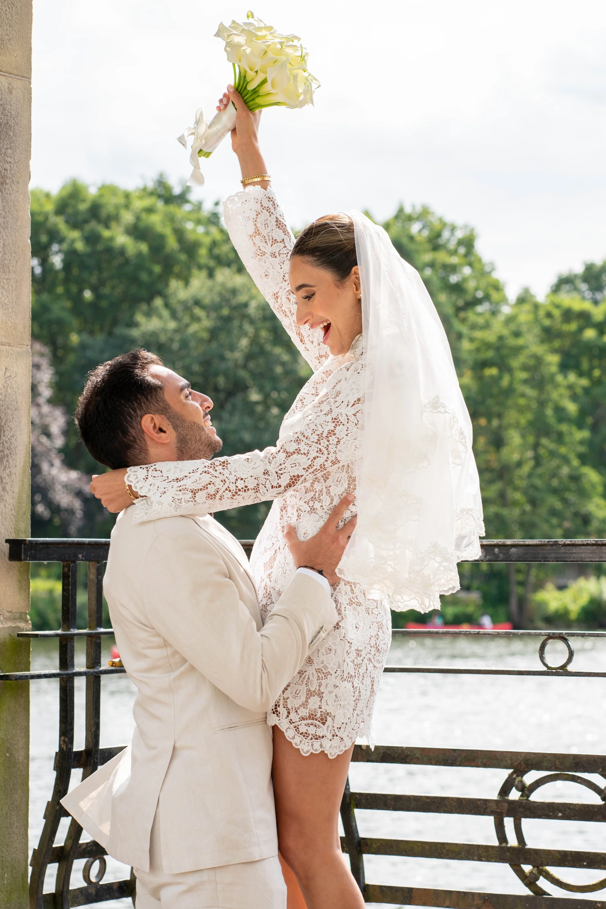 A happy couple celebrating their wedding outdoors near a body of water, with the woman holding a bouquet of white flowers and both dressed in white lace attire.