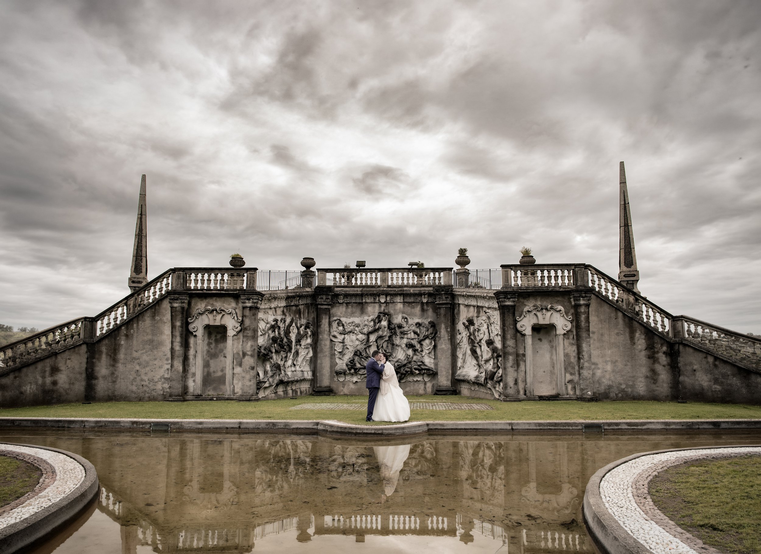 A bride and groom kissing in front of an ornate stone wall with sculptures and two tall obelisks on a cloudy day, reflected in a shallow water feature.