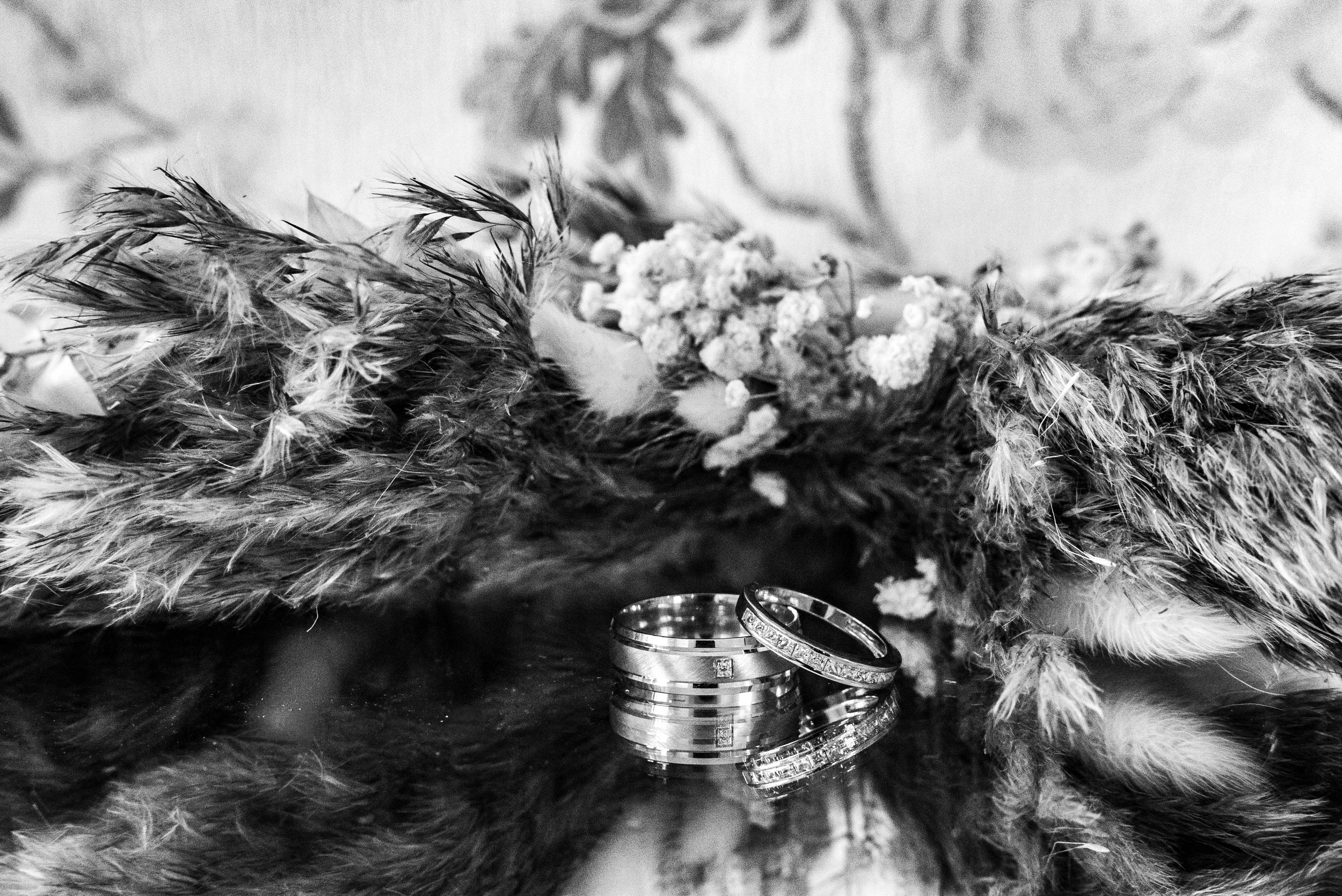 Close-up of wedding bands with a feathered flower arrangement in the background in black and white.