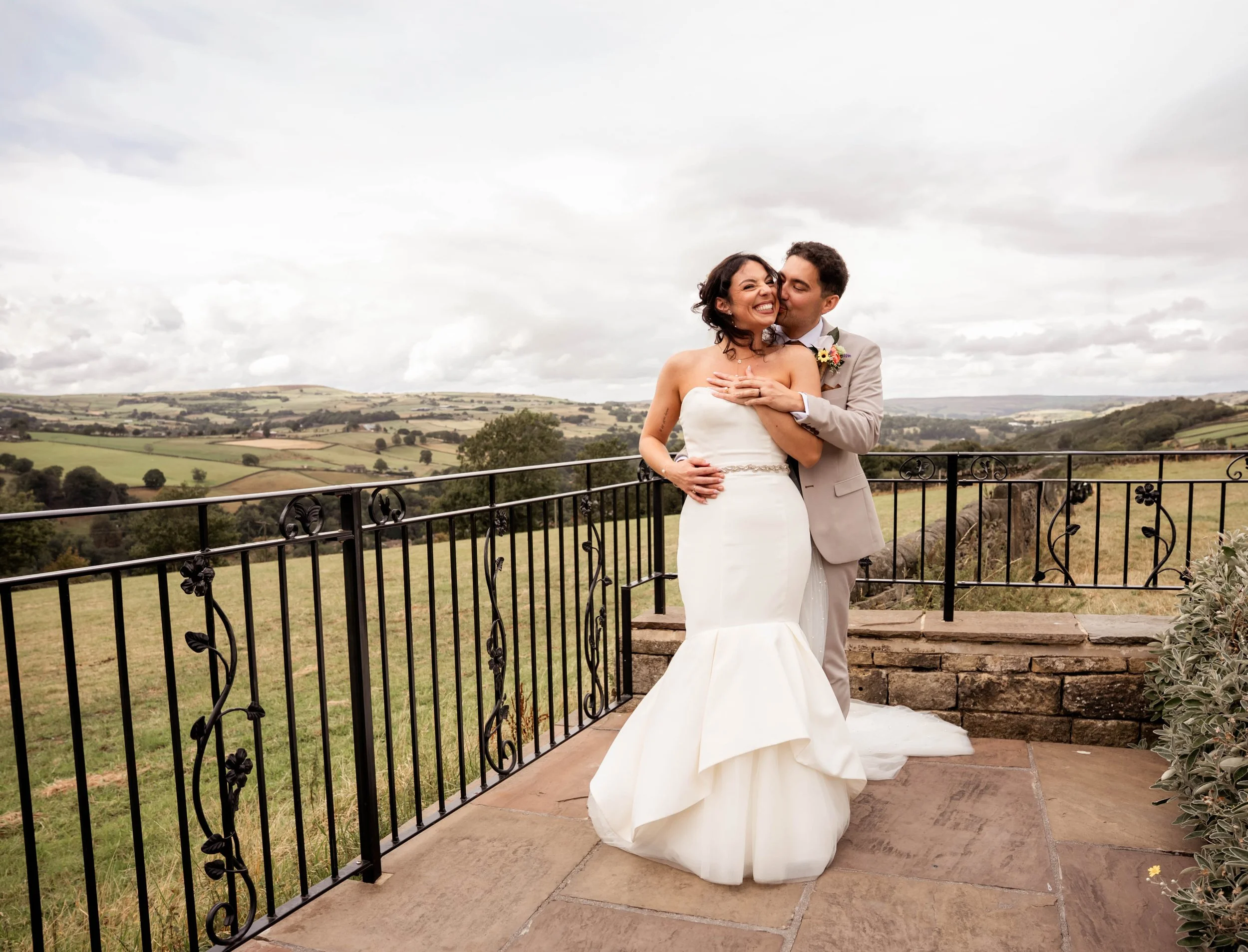 A bride and groom standing on a balcony overlooking a rural landscape with rolling hills under a cloudy sky. The groom is whispering to the bride, who is smiling. They are dressed in wedding attire, with the bride in a strapless white gown and the gr
