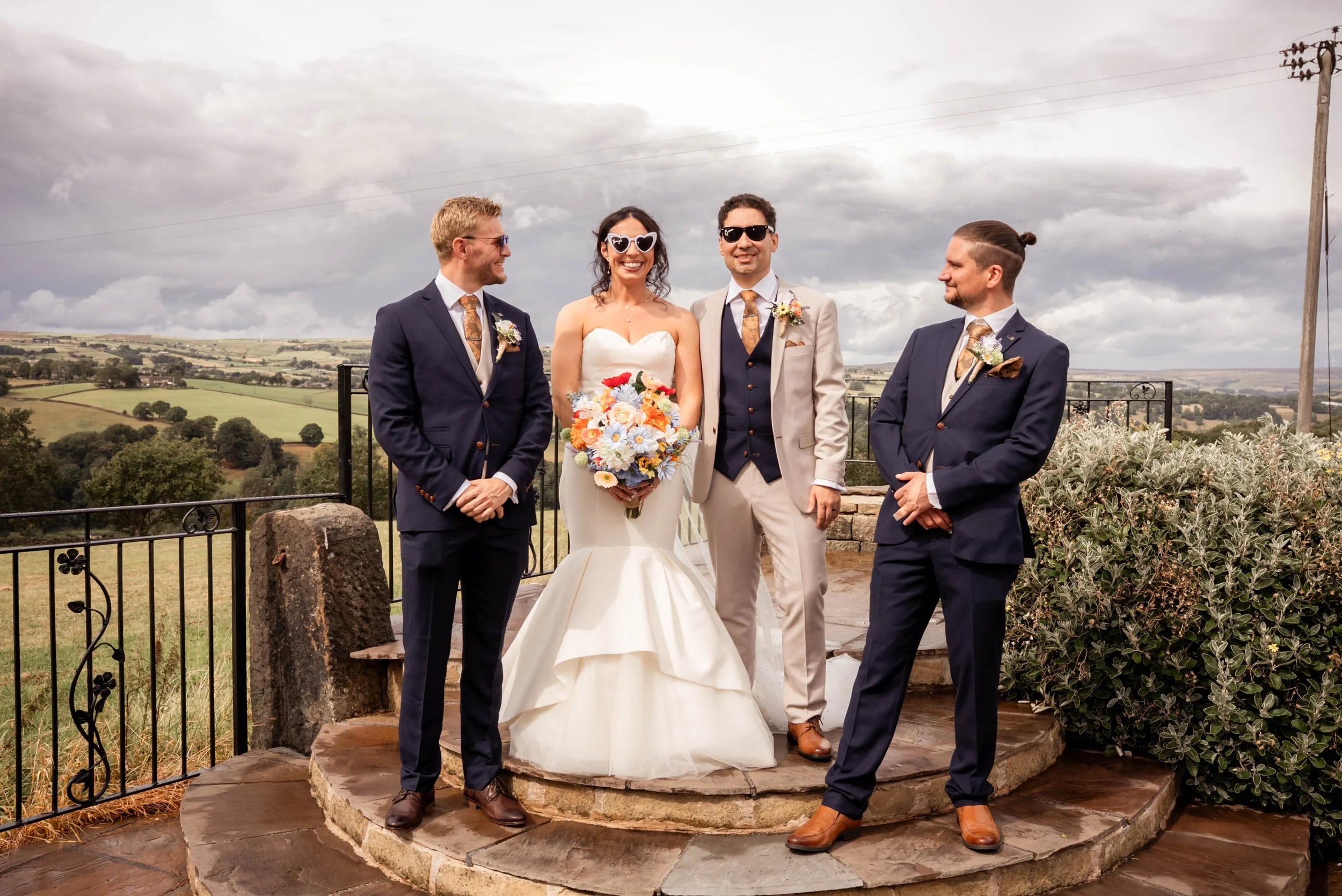 A wedding scene outdoors with five people: a bride in a white gown holding a colorful bouquet, two men and two women in suits, standing on a stone platform with a scenic landscape in the background under cloudy skies.