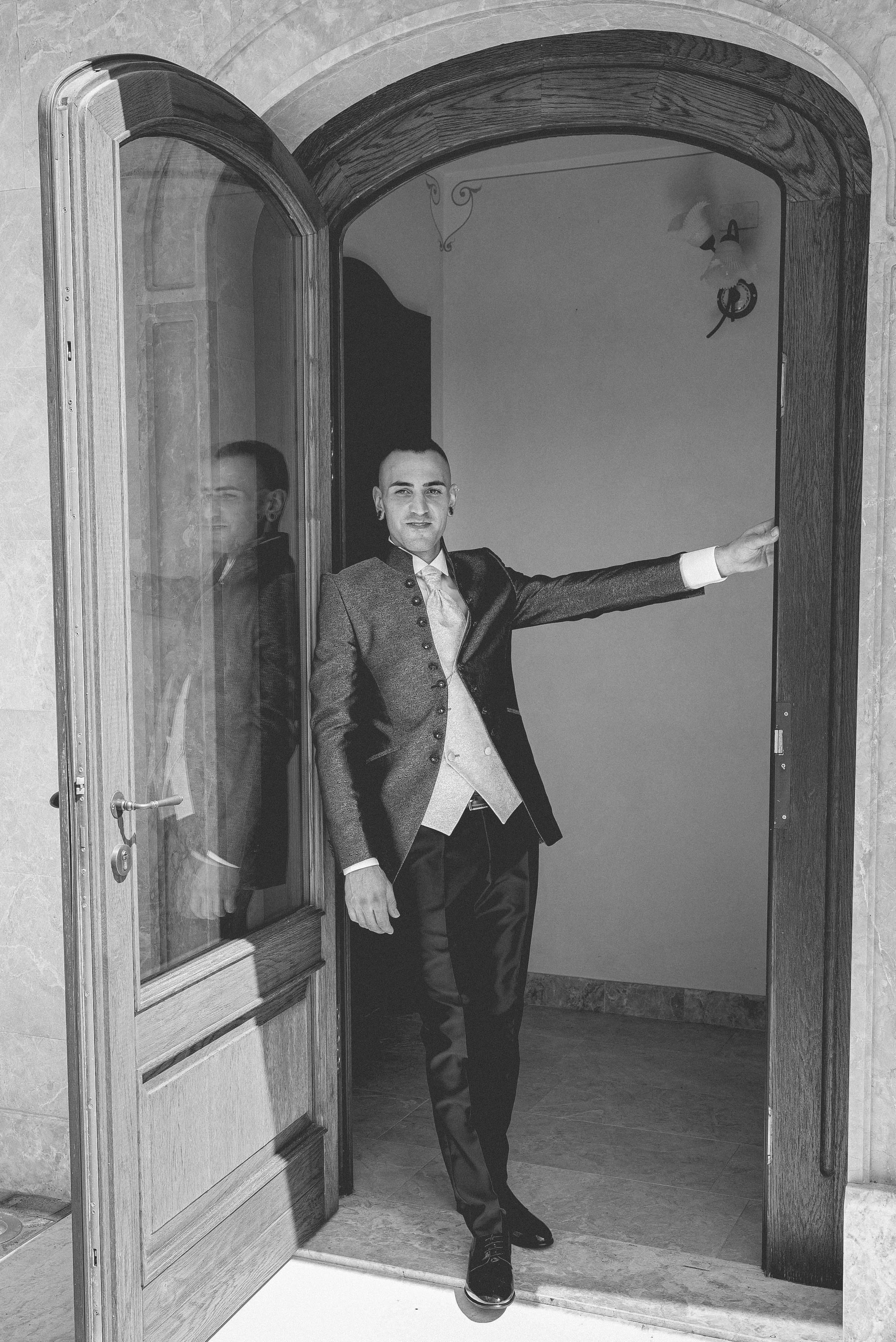 A person dressed in formal attire standing at a doorway, holding the door handle, with a reflection of themselves visible in the glass of the door.