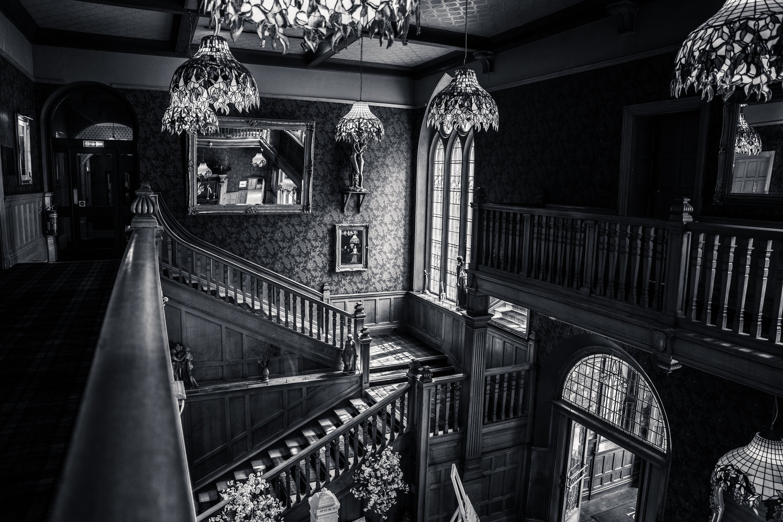 Interior view of an elegant, dark-themed staircase hall with ornate chandeliers hanging from the ceiling, framed mirrors, and decorative statues, with large arched windows allowing natural light.