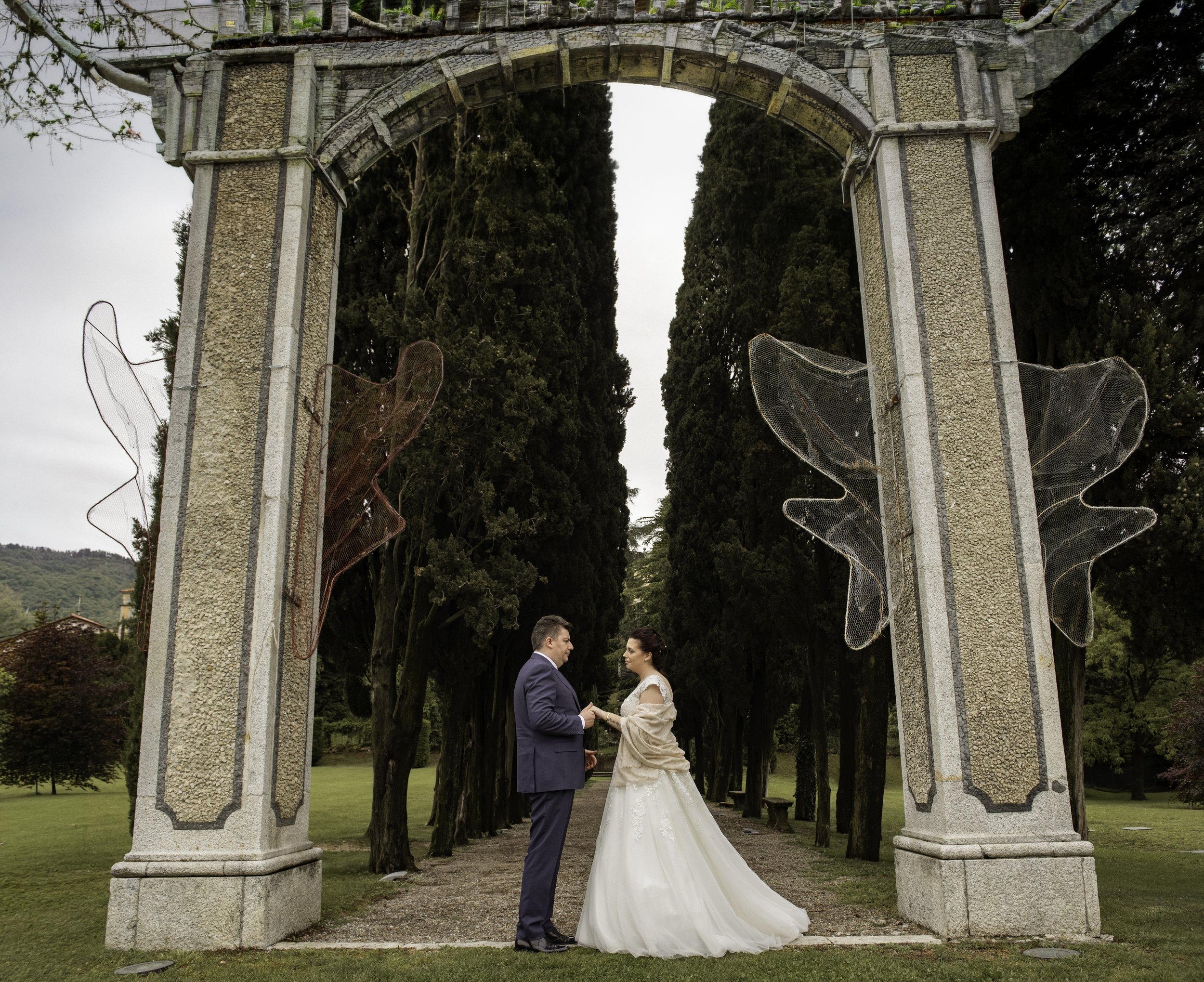 A bride and groom holding hands under a stone archway with tall trees in the background during a wedding ceremony.