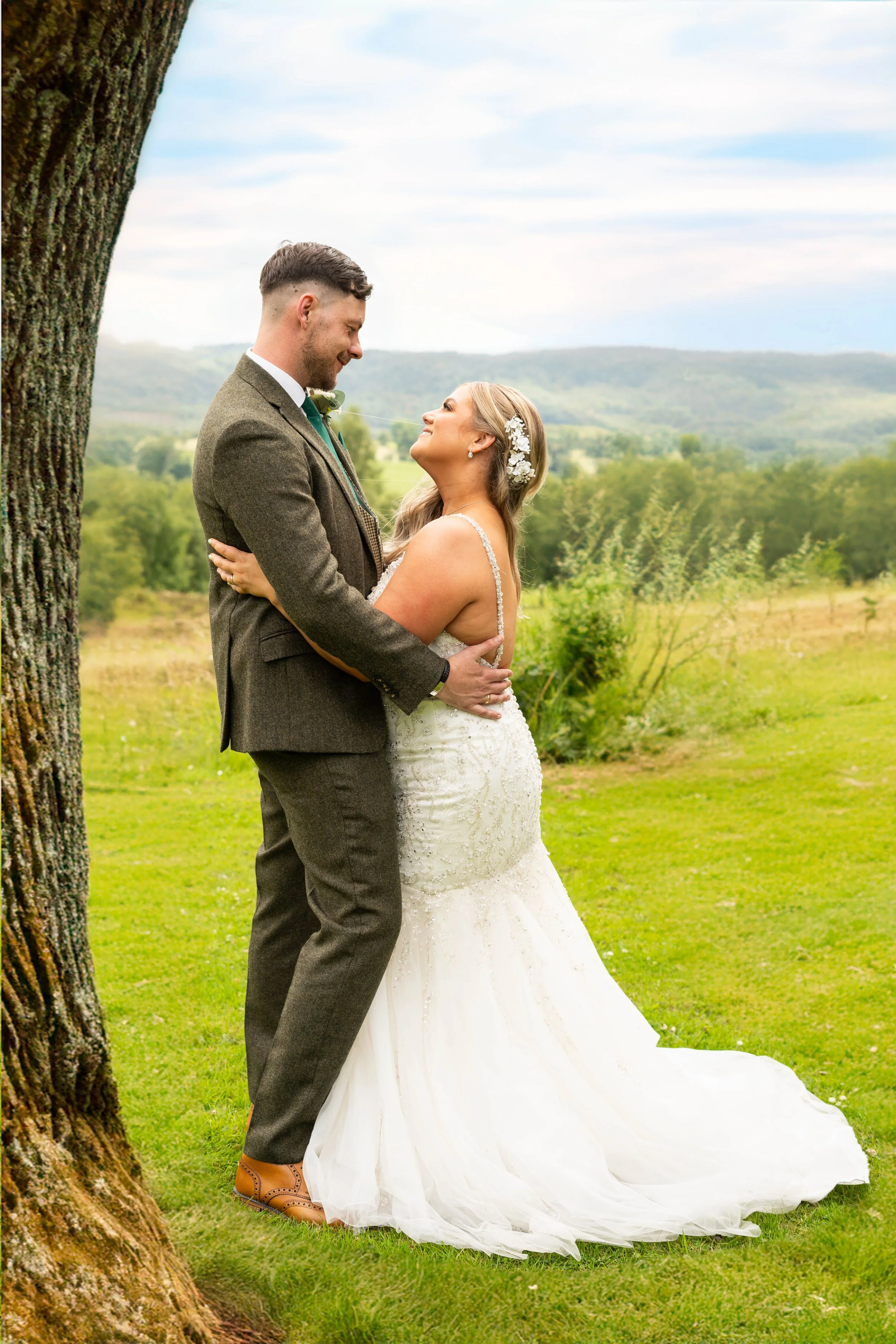 A bride and groom stand outdoors on green grass, gazing into each other's eyes, with a scenic landscape of rolling hills and trees in the background. The bride wears a white lace wedding gown with floral hair accessories, and the groom wears a brown