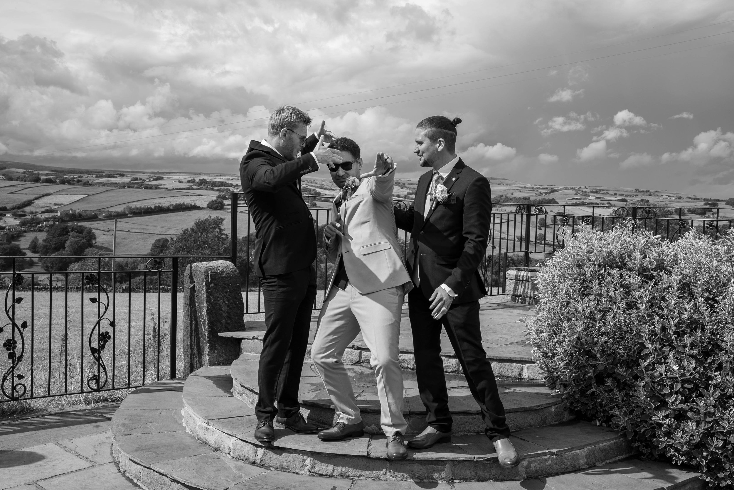 Three men in suits on a stone platform with a scenic countryside background, one of whom is wearing sunglasses, one is gesturing with his hands, and the third has a man bun, engaged in a conversation or celebration.