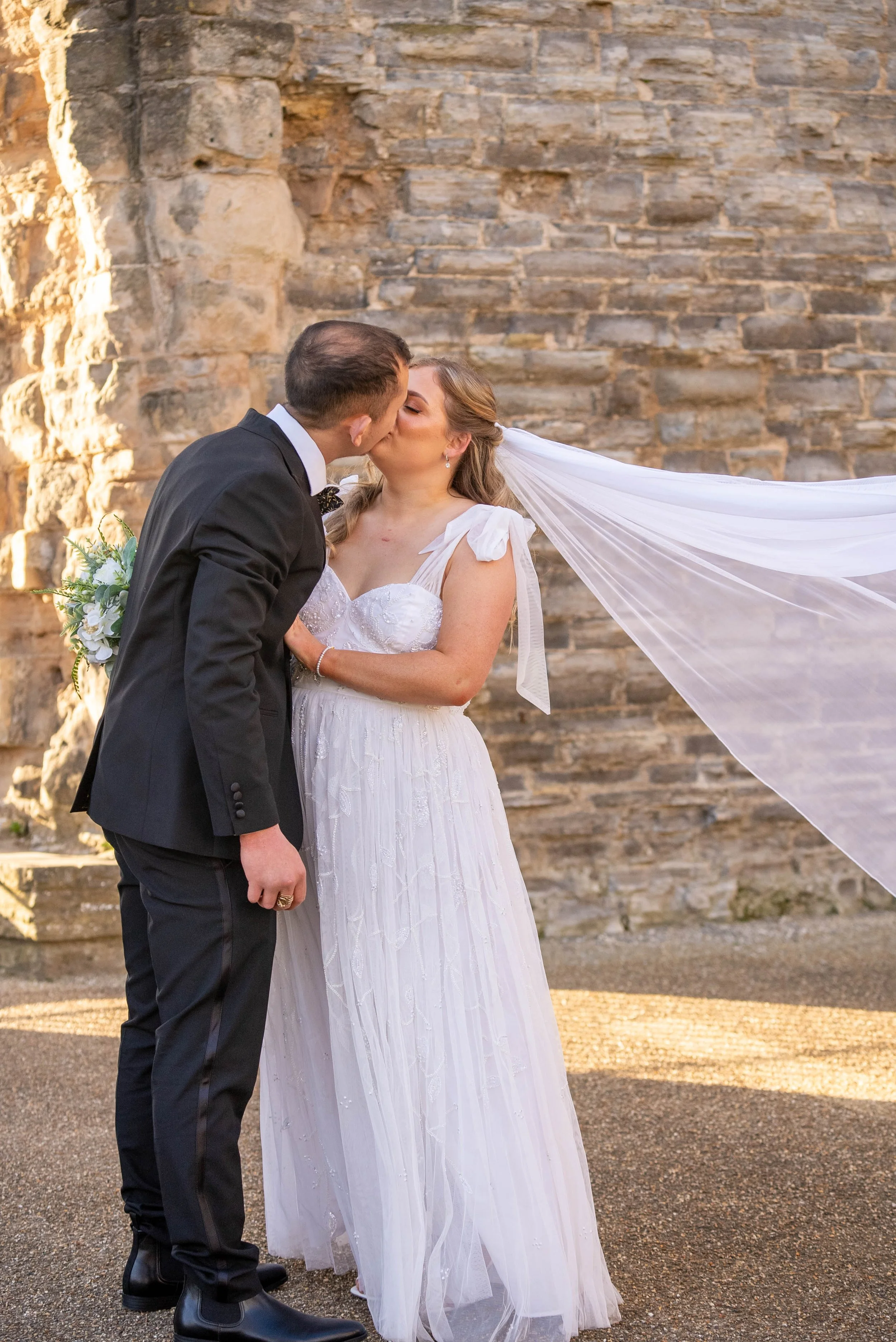 A bride and groom sharing a kiss outdoors during their wedding ceremony, with the bride wearing a white wedding dress and veil, and the groom in a black suit with a bow tie, against a stone wall background.