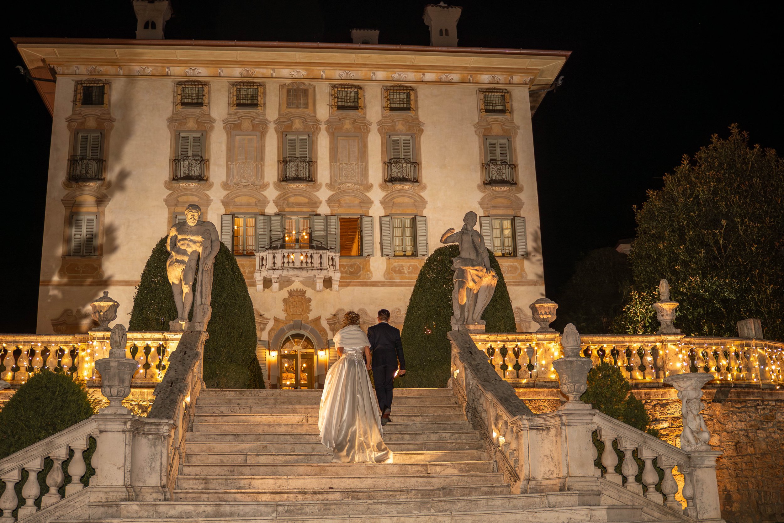 A nighttime scene of a grand, historic building illuminated with warm lighting, featuring ornate architectural details and iron balconies. In the foreground, a staircase with sculptures of seated figures and decorative railings leads up to the entran