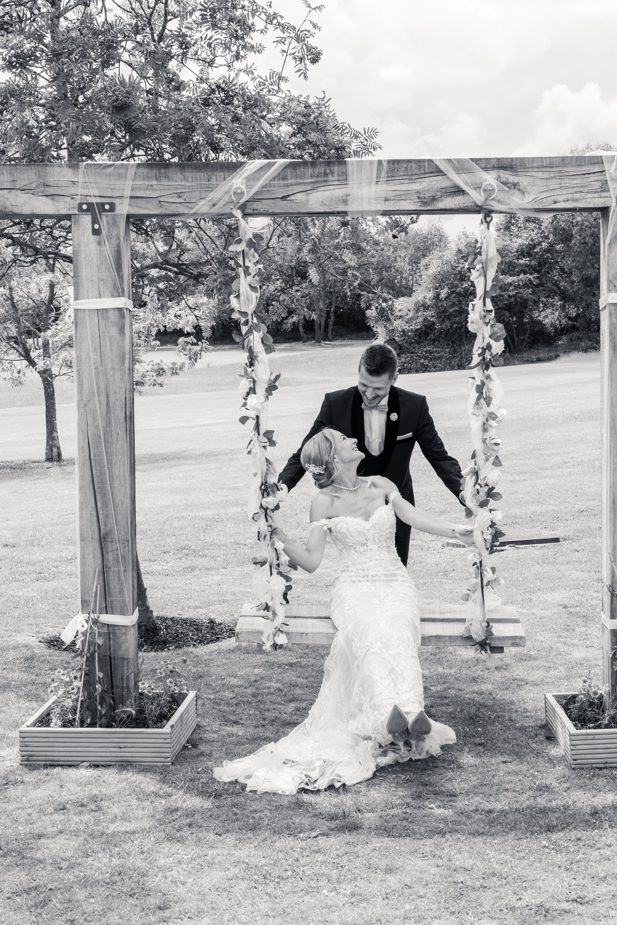 A bride and groom at an outdoor wedding ceremony under a wooden arch decorated with vines and flowers, with trees and cloudy sky in the background.
