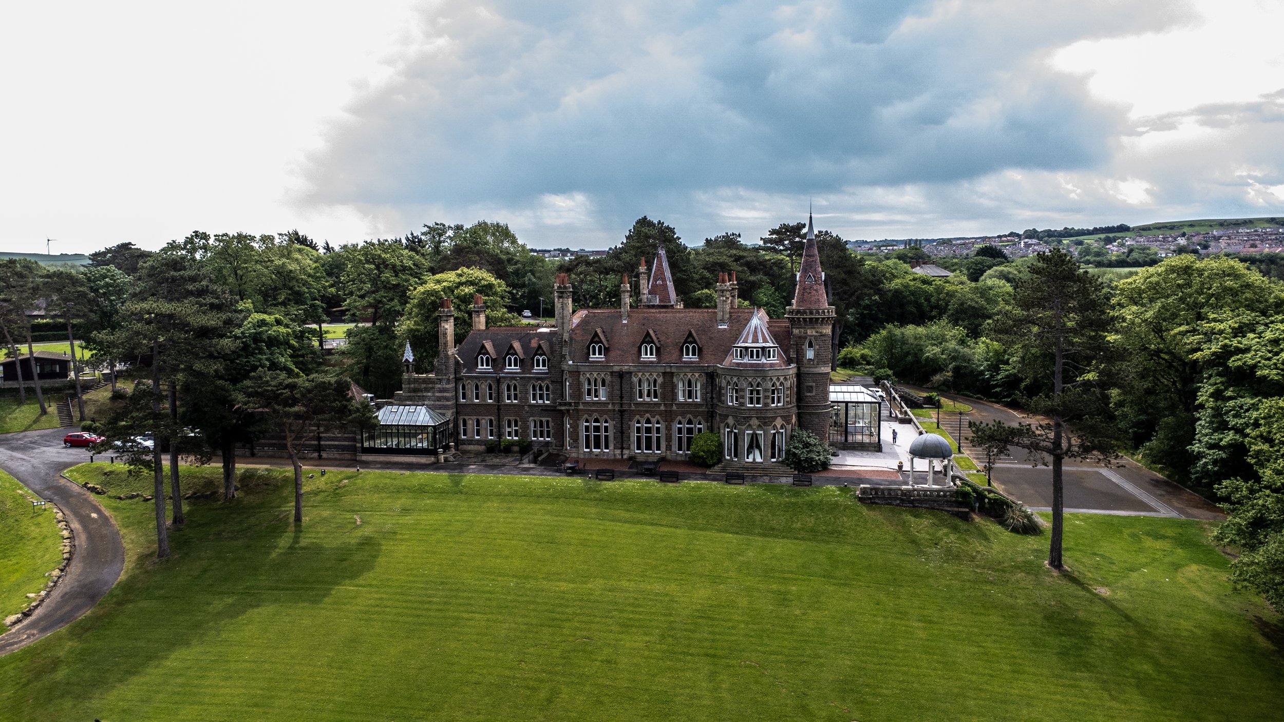 Historic castle-like building with pointed towers, situated amid lush green trees and an open lawn, under a cloudy sky.