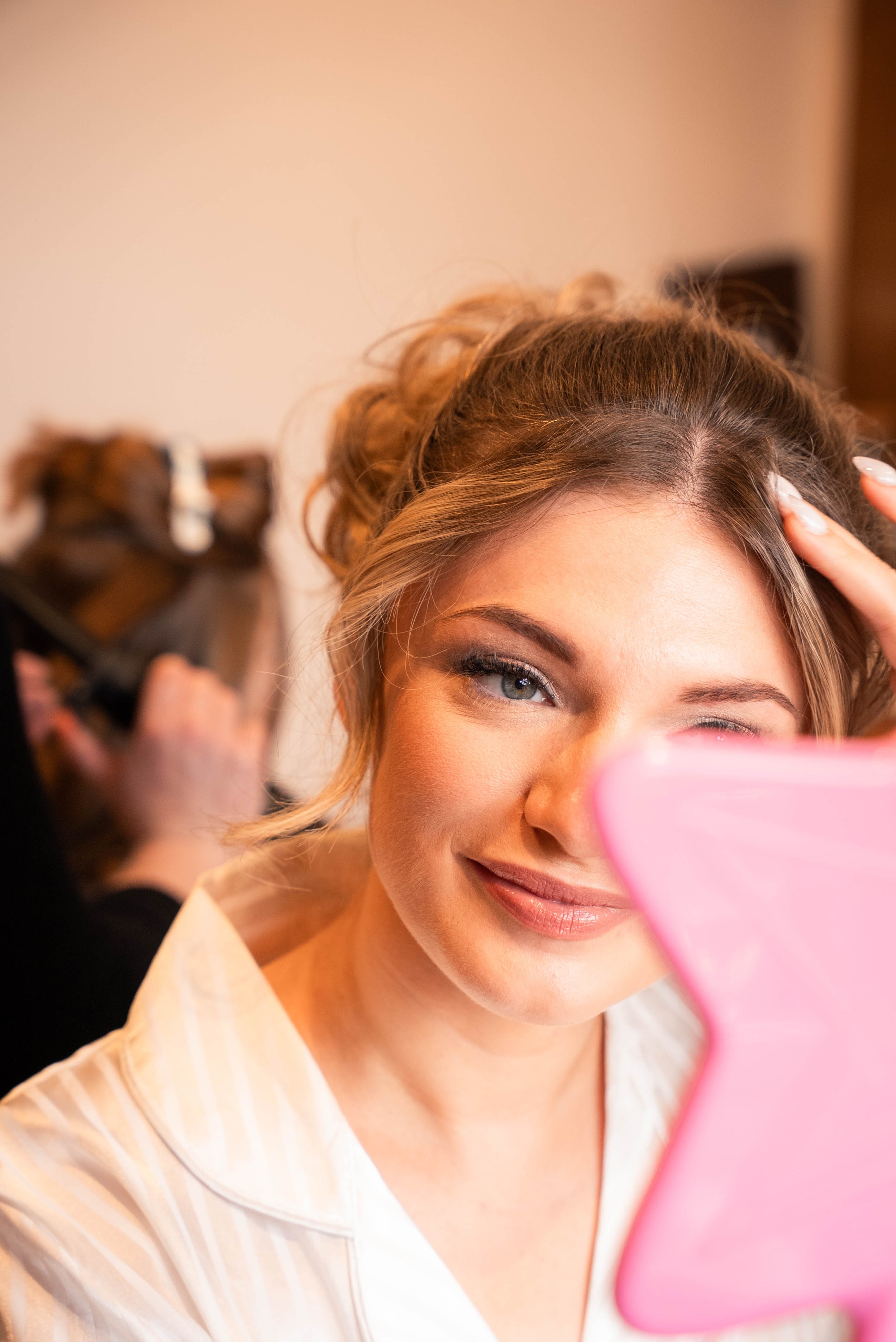 A woman with styled hair is looking into a pink mirror, smiling, as a hairstylist works on her hair in the background.