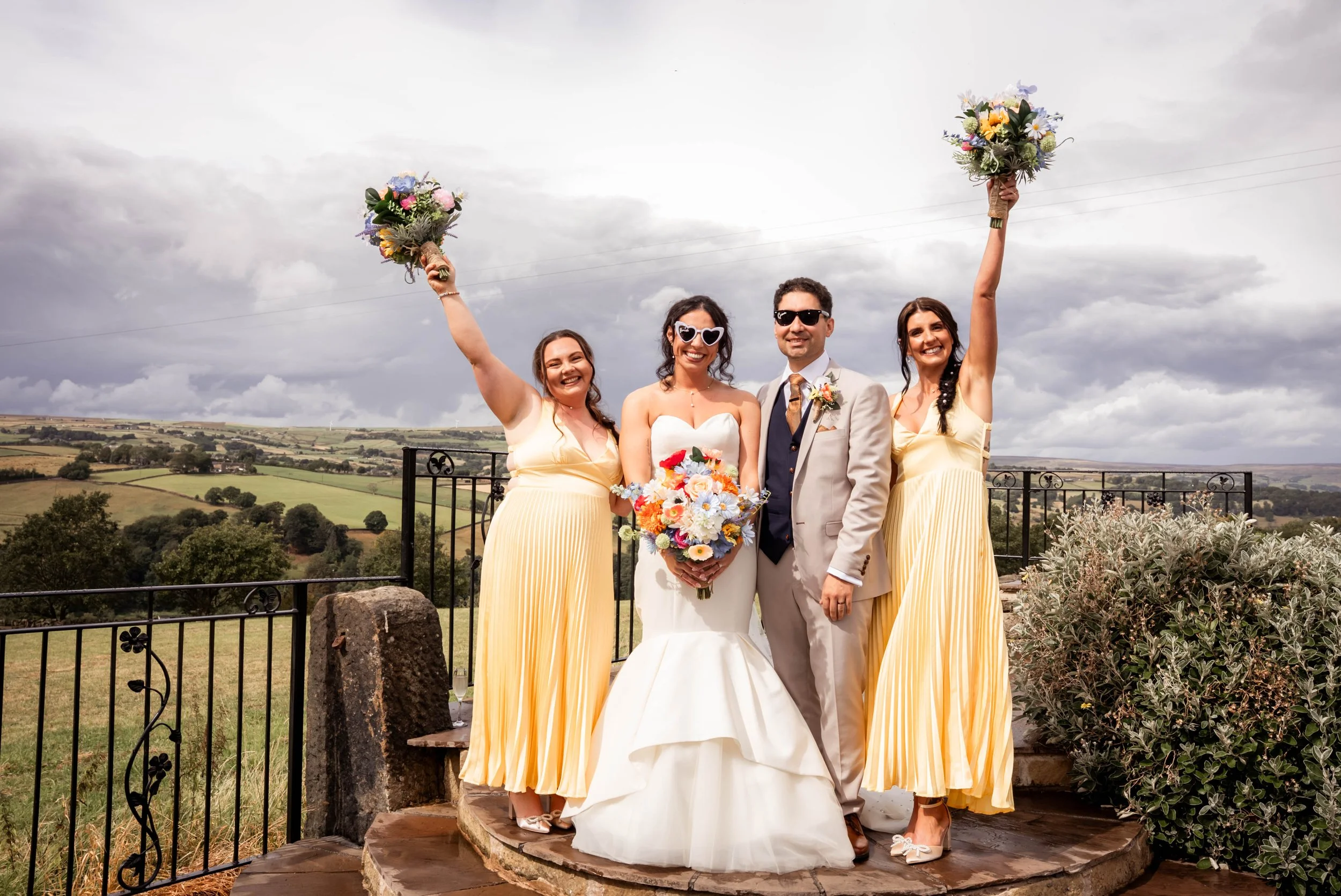 A group of five people at a wedding outdoors, with a scenic landscape and cloudy sky behind them. The bride and groom stand in the center, with the bride holding a large bouquet and wearing a white wedding dress. The groom wears a light gray suit and