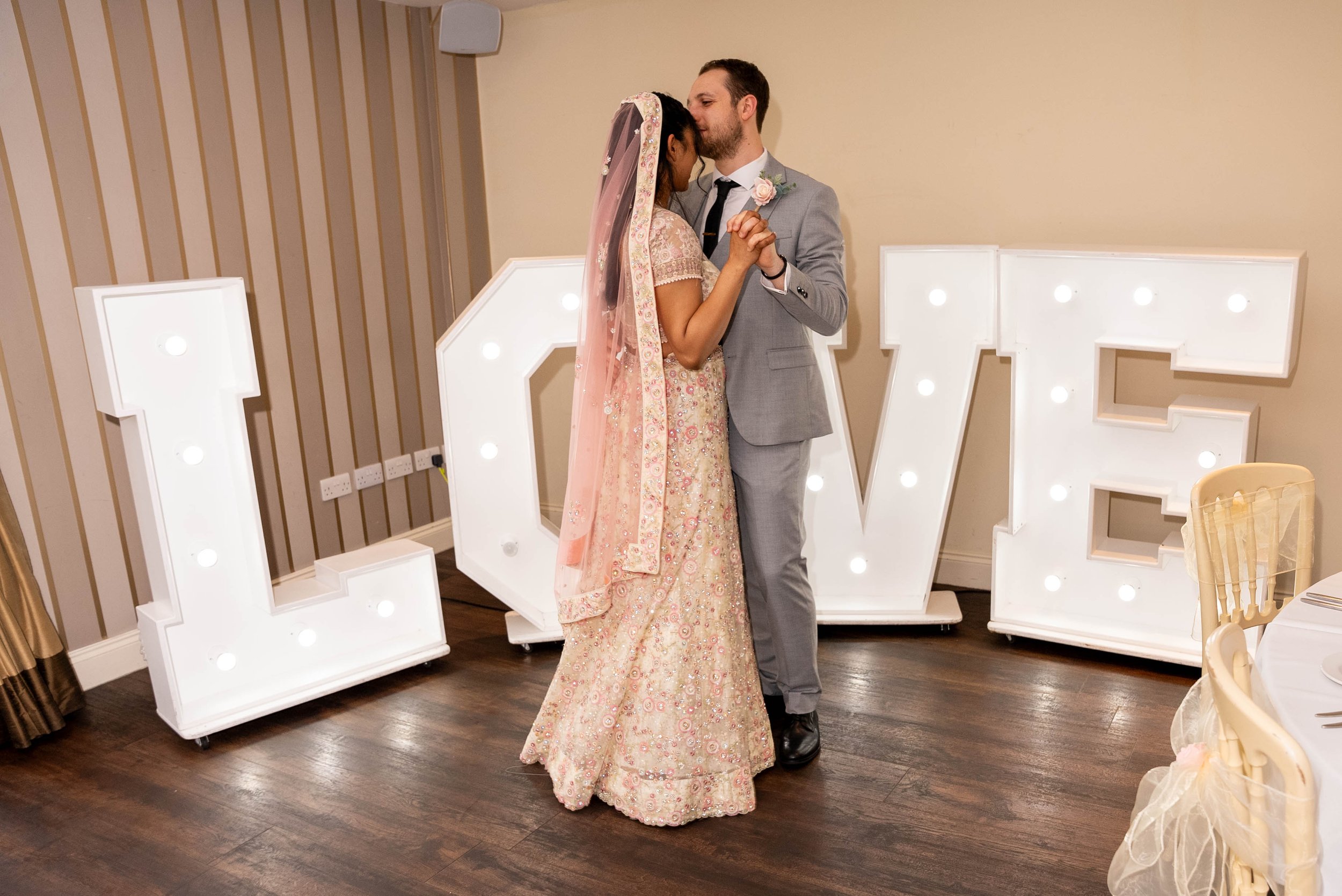 A bride and groom share a first dance in front of large illuminated letters spelling 'LOVE' at a wedding reception.