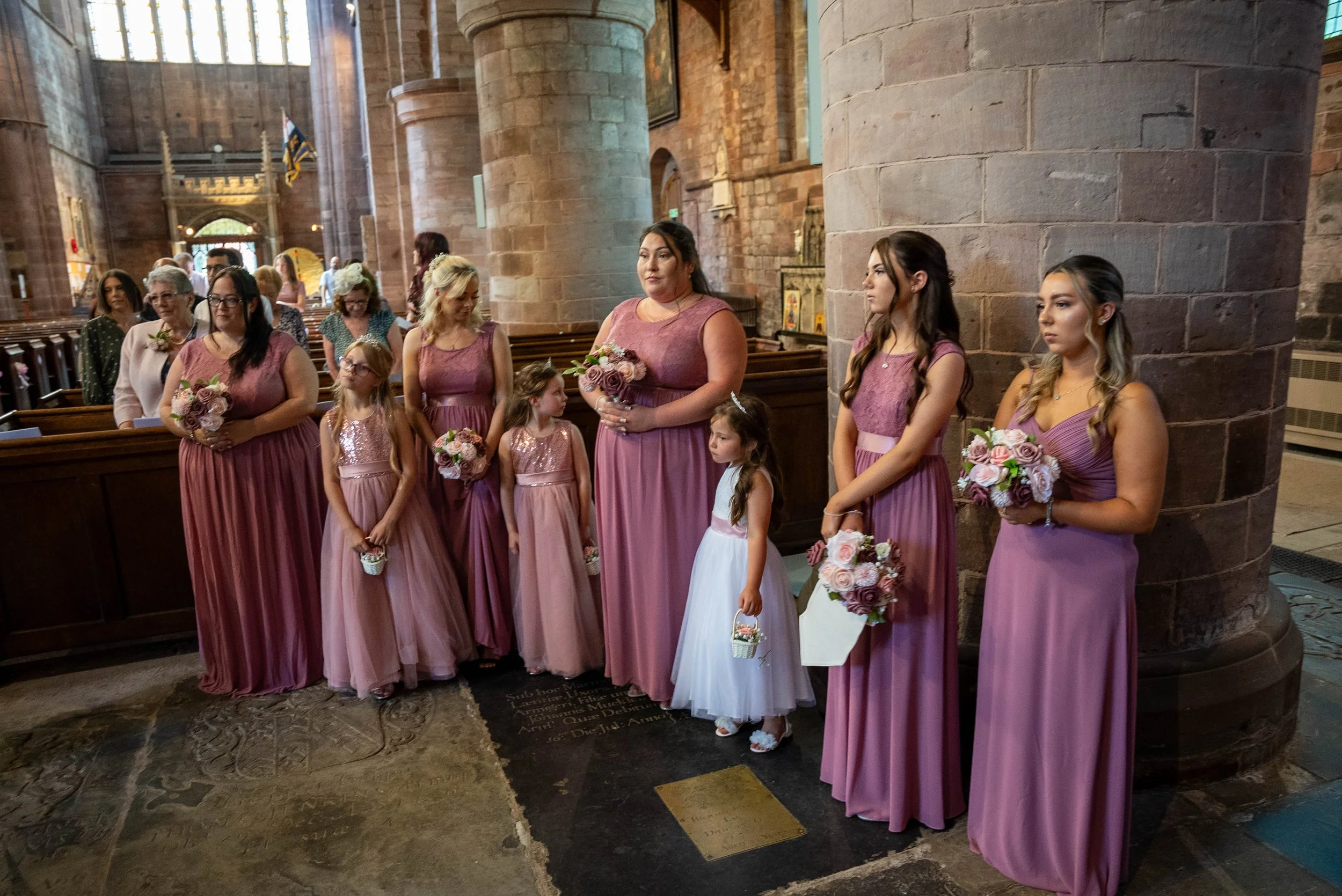 A group of women and young girls dressed in pink and white dresses standing inside a church. They are holding bouquets and small baskets, likely participating in a wedding ceremony.