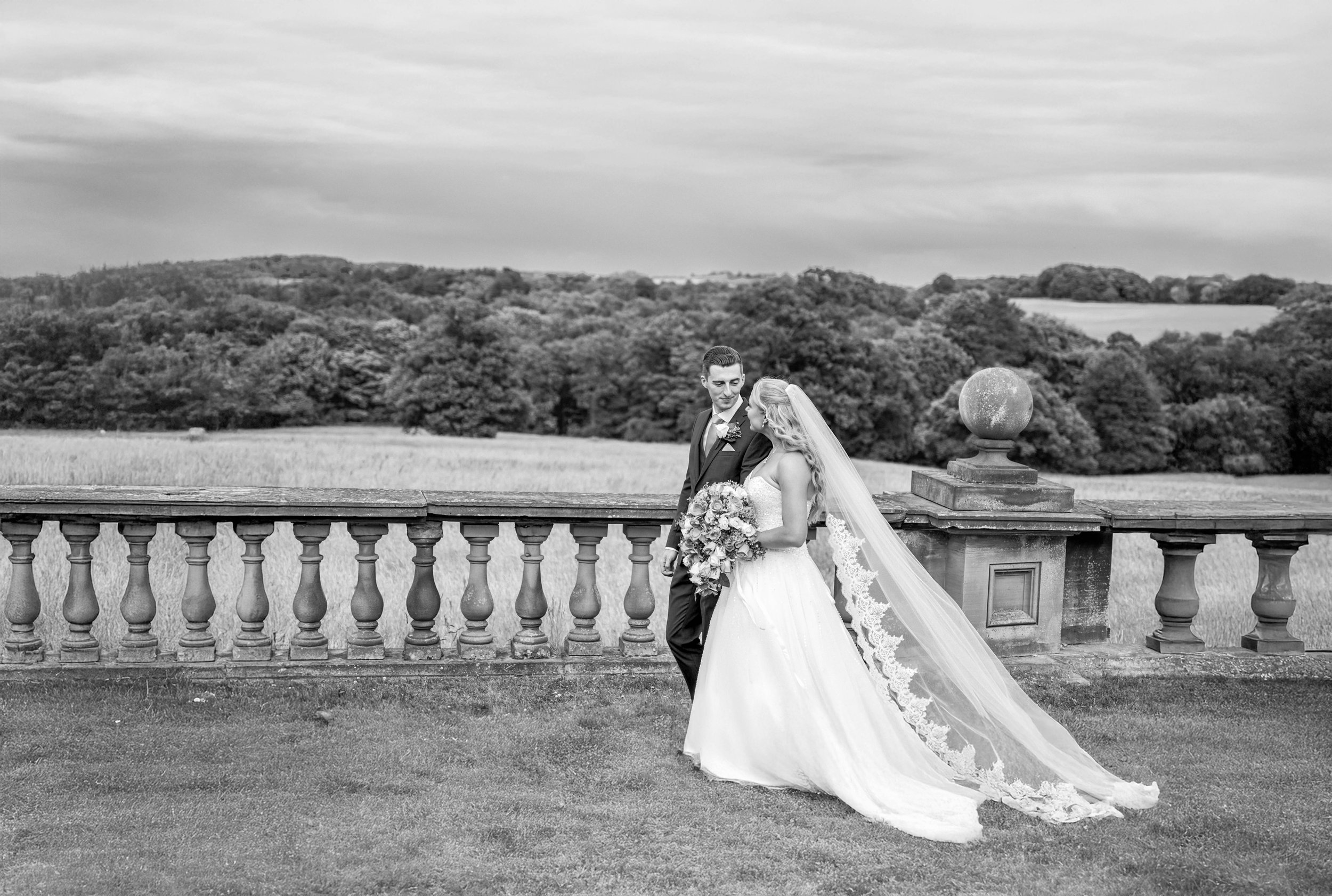 Black and white photo of a bride and groom standing close together on a grassy area with a stone balustrade, overlooking a landscape with trees and rolling hills. The bride is holding a large bouquet and wearing a long veil.