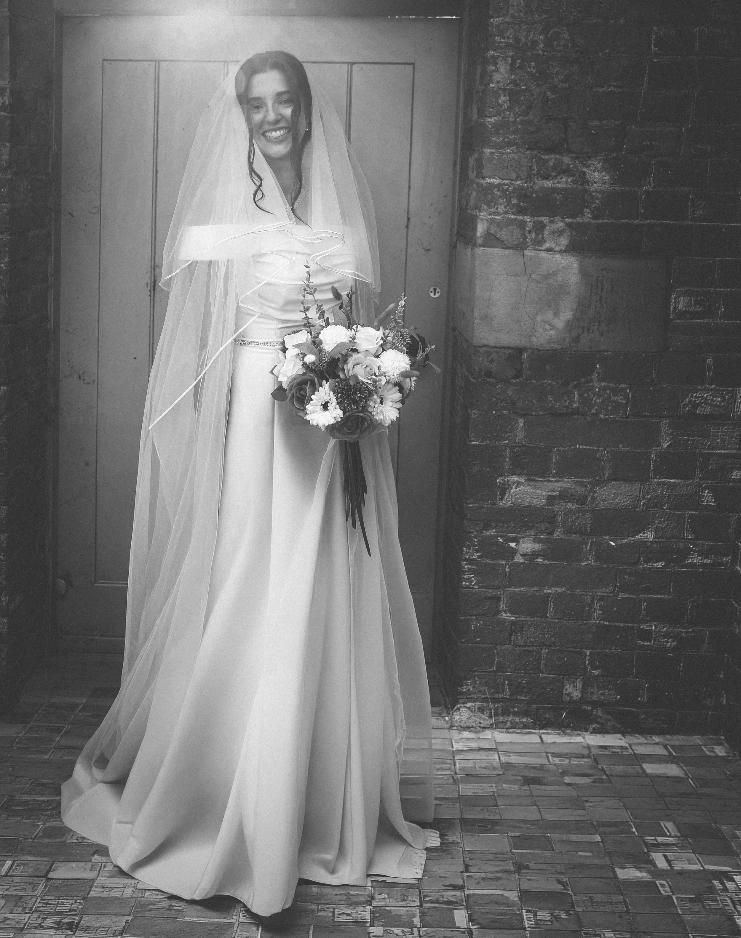 A woman dressed in a wedding gown and veil, holding a bouquet of flowers, standing in a corridor with brick walls.