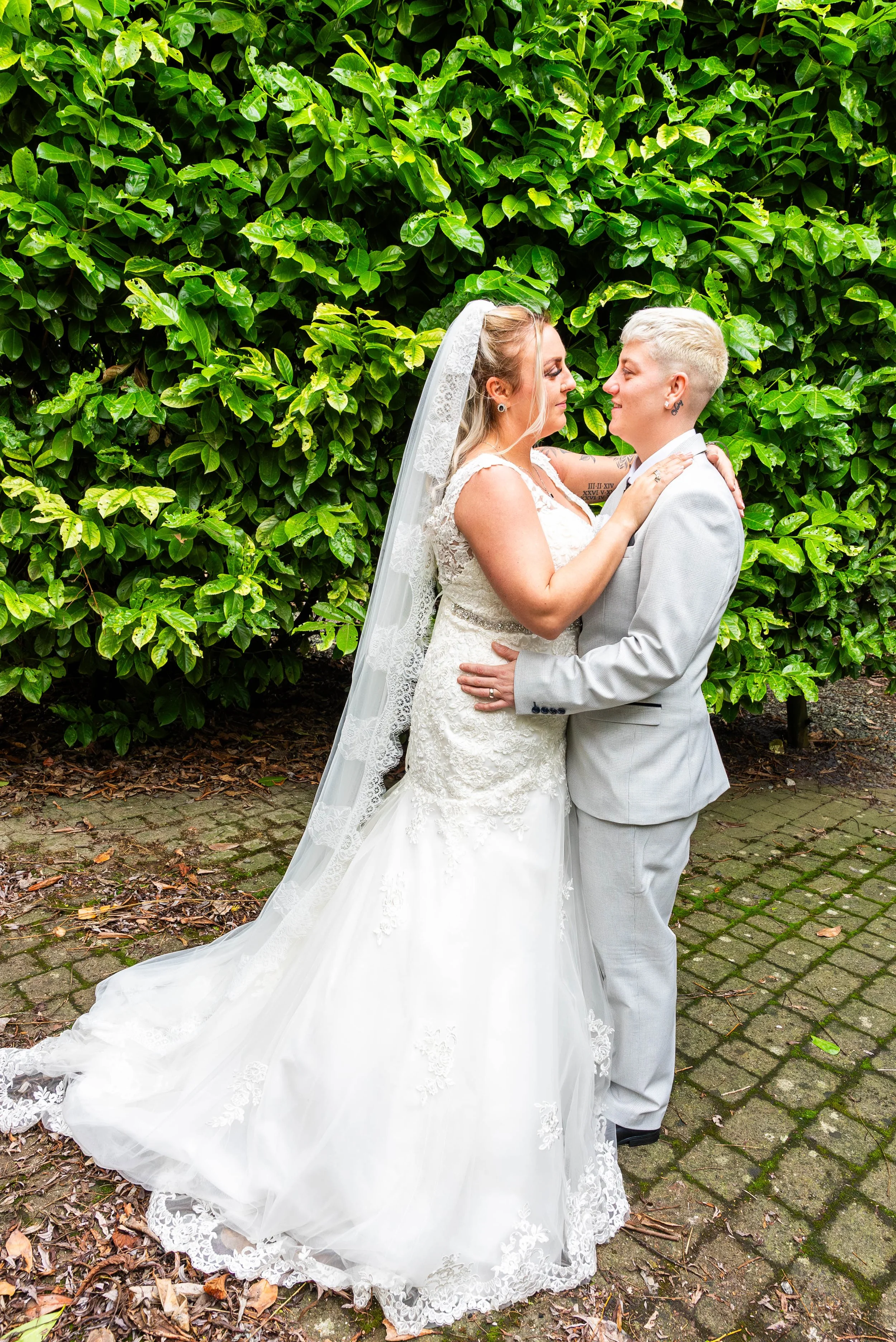 Two women in wedding attire embrace and look into each other's eyes in front of a green leafy bush, standing on a cobblestone path.