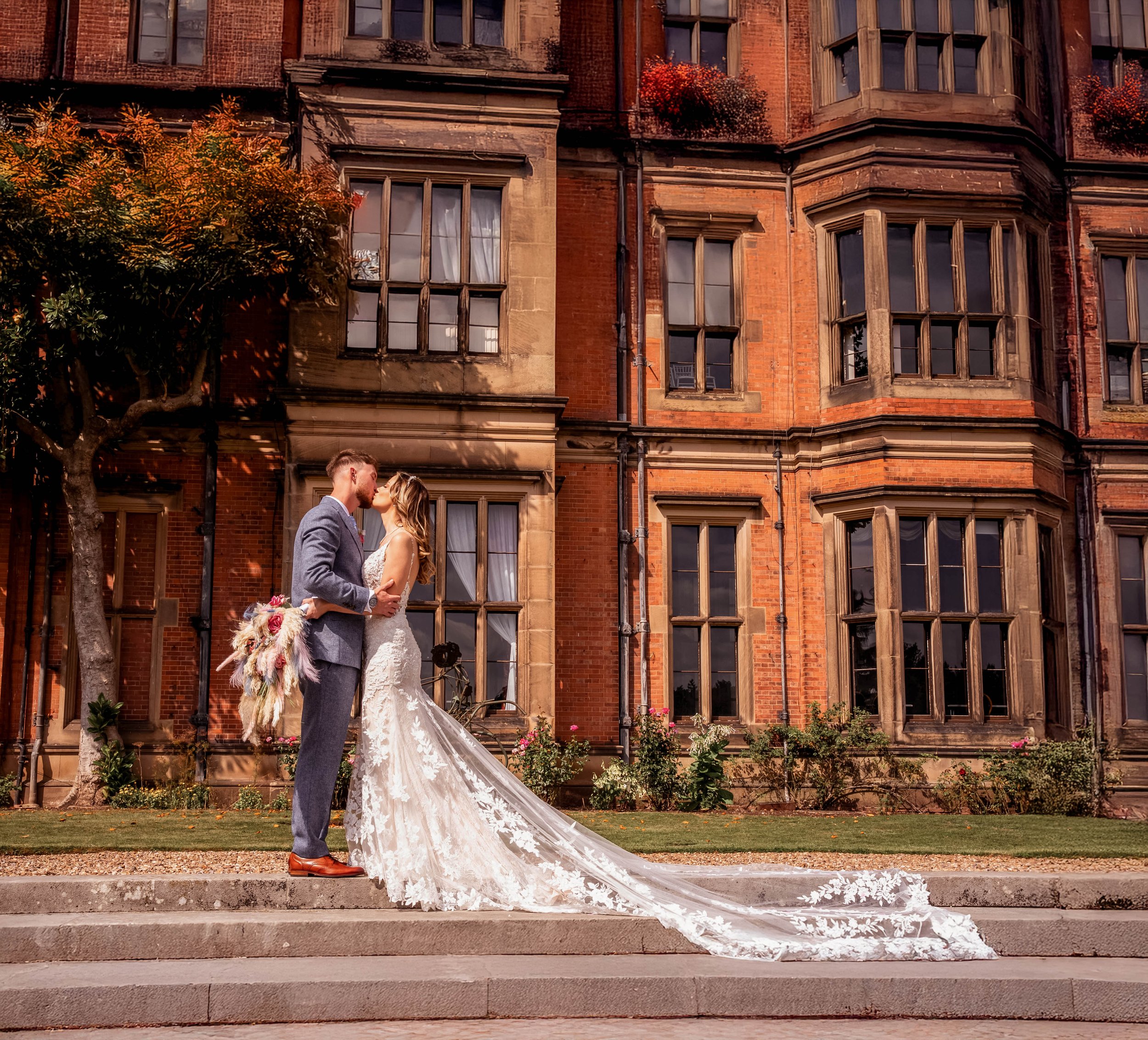 A bride and groom share a kiss on steps in front of a historic brick building, with the bride wearing a lace wedding dress and the groom in a suit. The bride holds a bouquet of flowers, and the couple is surrounded by trees and flower beds.