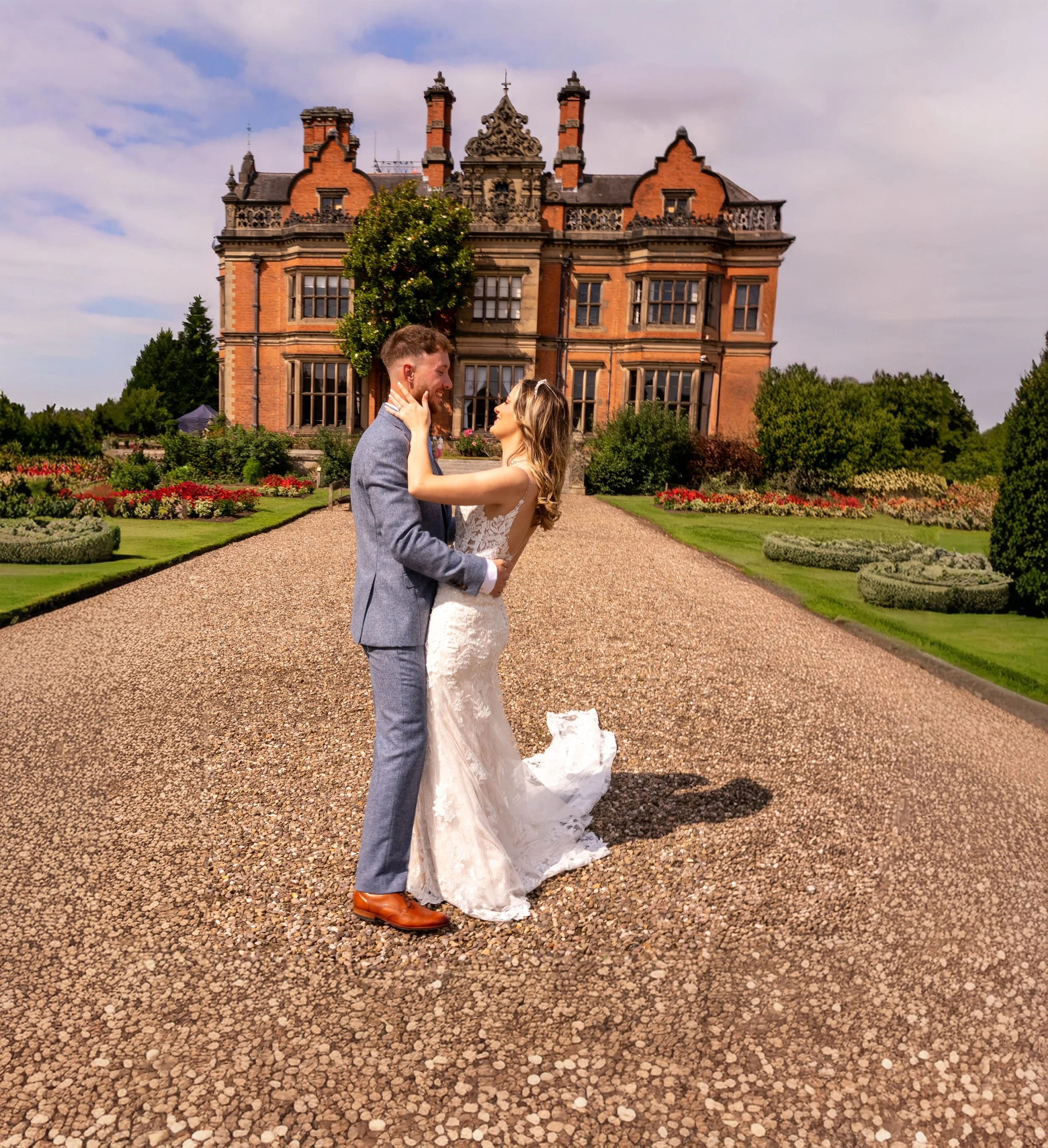A bride and groom are standing on a gravel driveway in front of a large, historic red-brick mansion with ornate architectural details. The couple is smiling and looking into each other's eyes, with the bride holding the groom's face and the groom gen