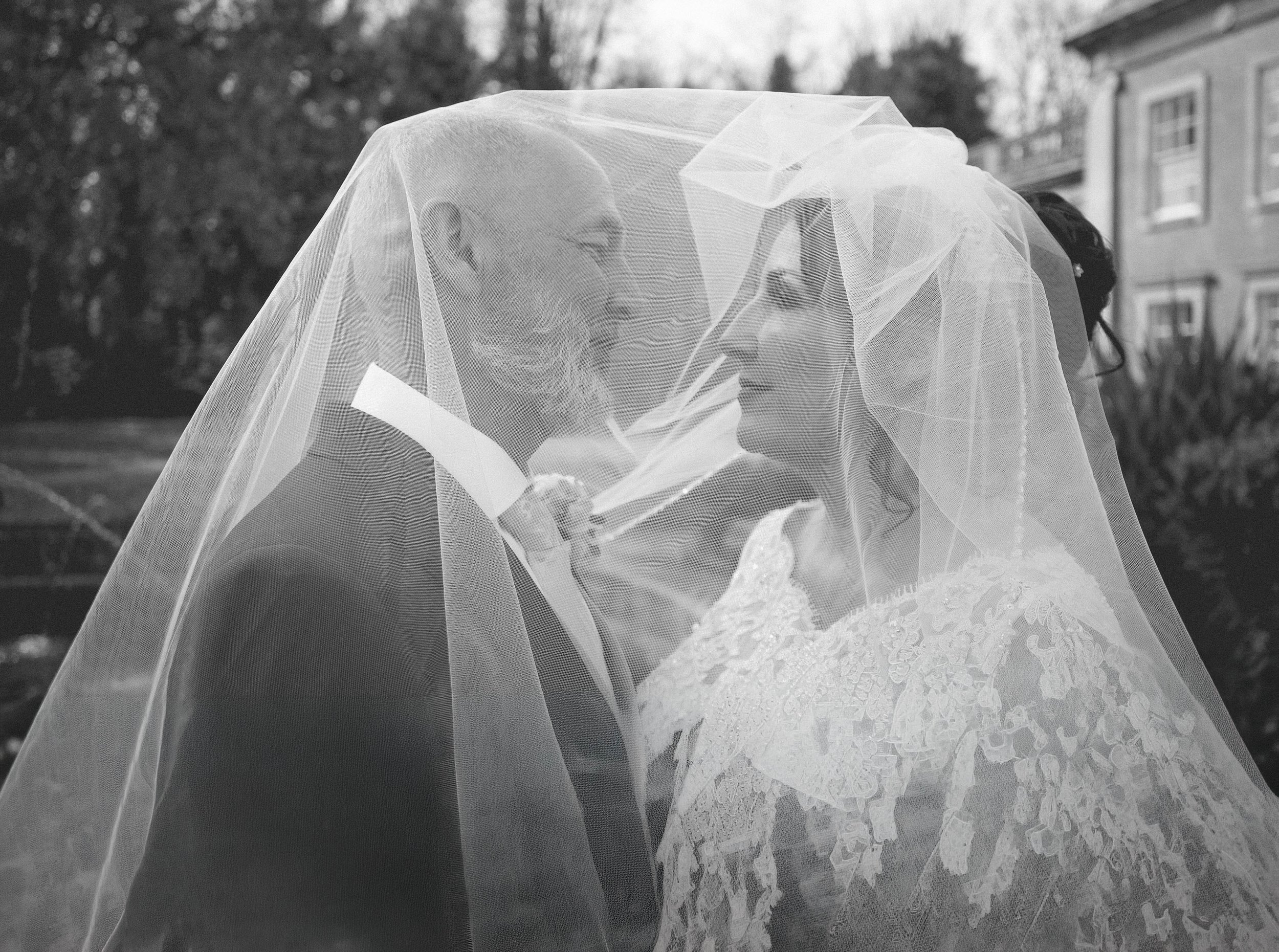 Black and white photo of a bride and groom standing close together, with a veil covering both of their faces, outdoors.