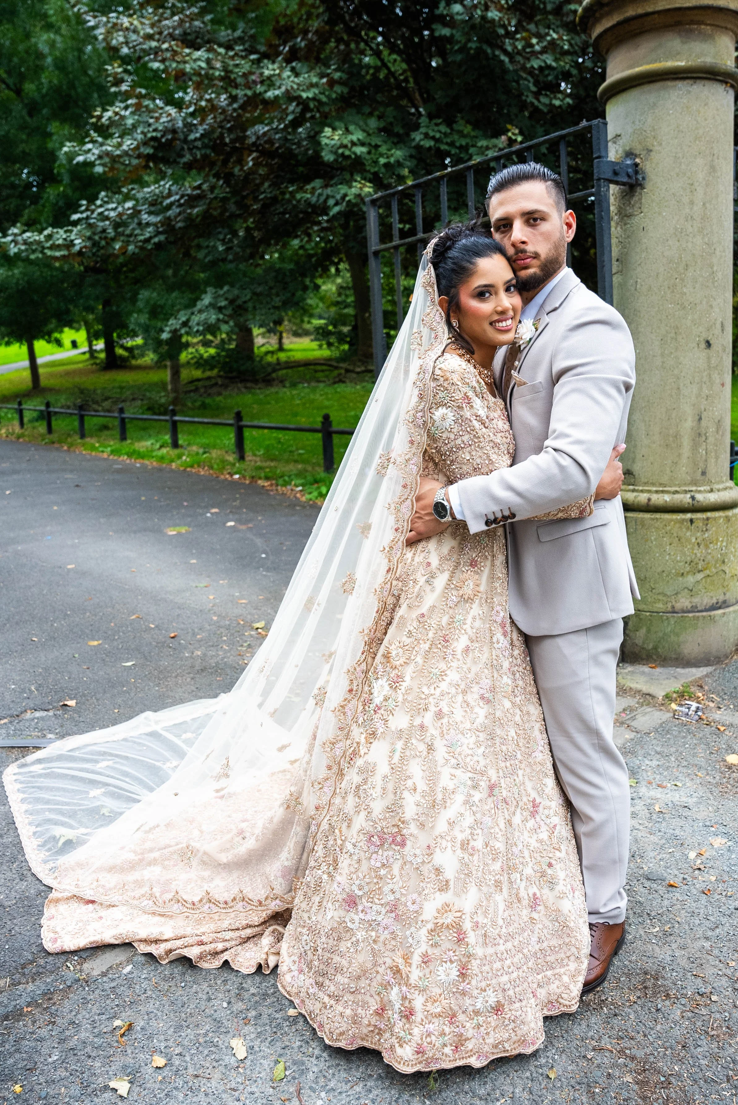 A newlywed couple embraces outdoors in front of a stone pillar and a black iron fence, surrounded by trees and greenery. The bride wears an ornate, beaded gown with a long veil, and the groom is dressed in a light gray suit.
