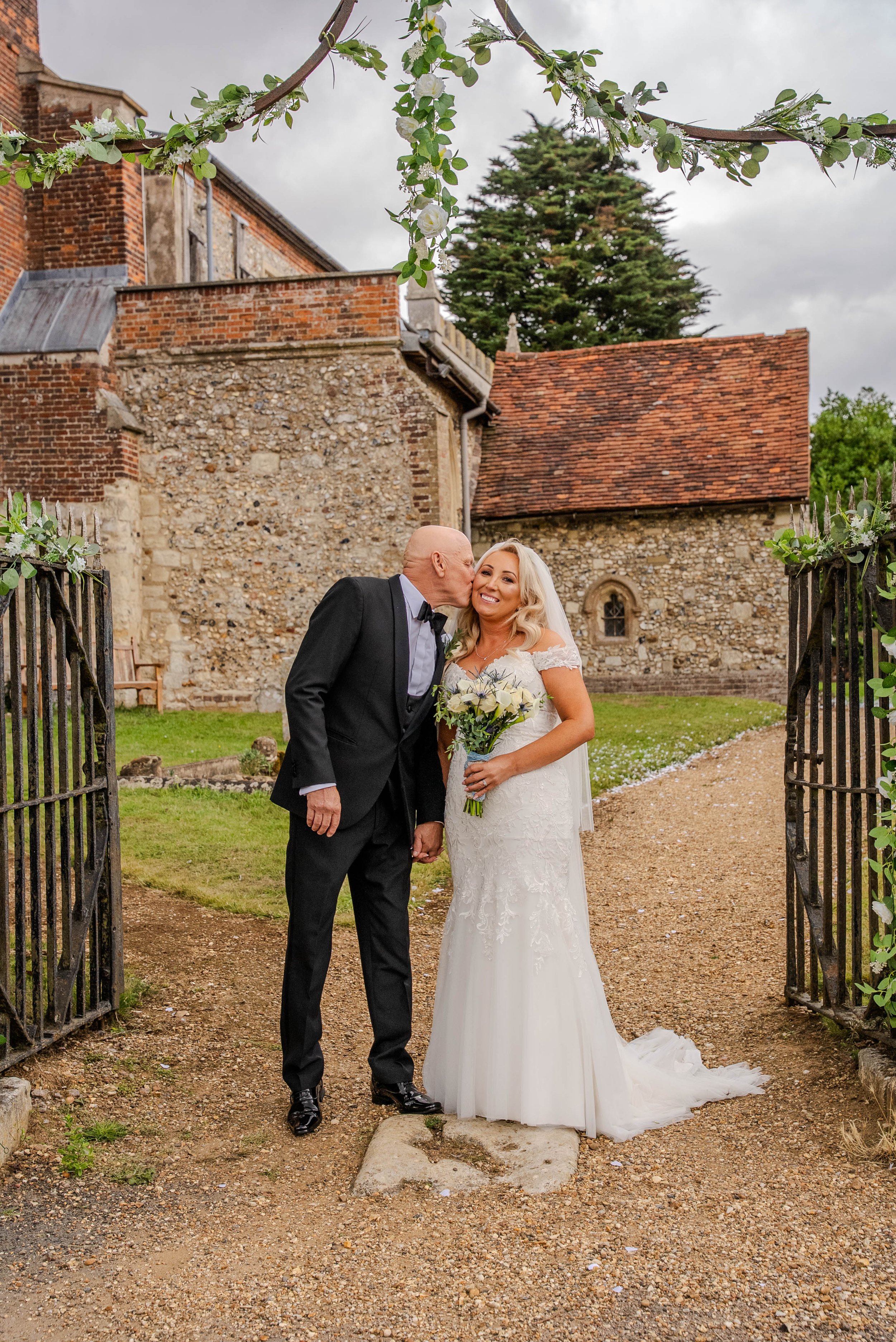 A bride in a white wedding dress holding a bouquet of flowers, smiling as a man in a black tuxedo kisses her cheek, standing at the entrance of a historic stone building with a brick roof, decorated with greenery on the iron gates.