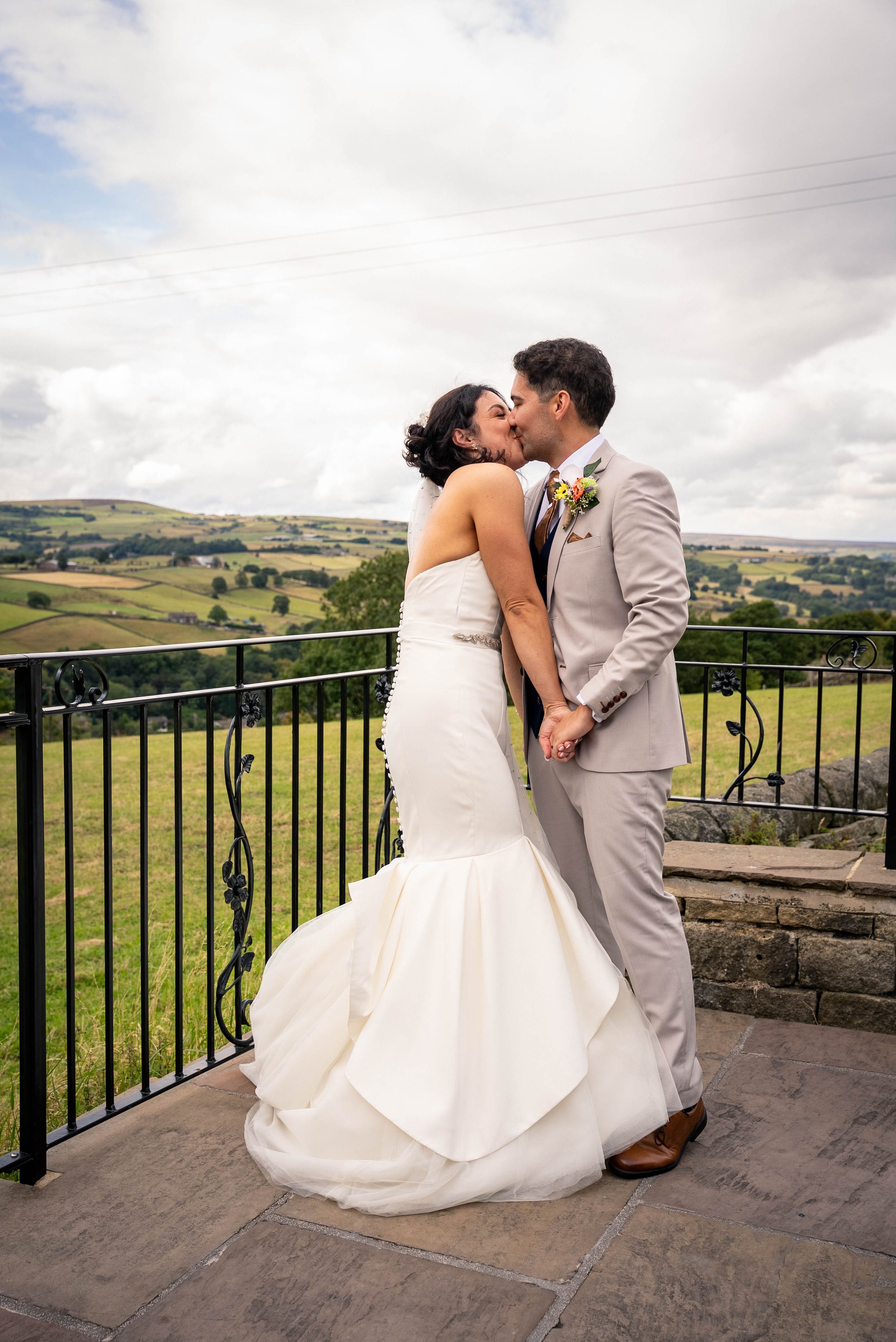 A newlywed couple sharing a kiss on a balcony overlooking a green landscape with rolling hills.
