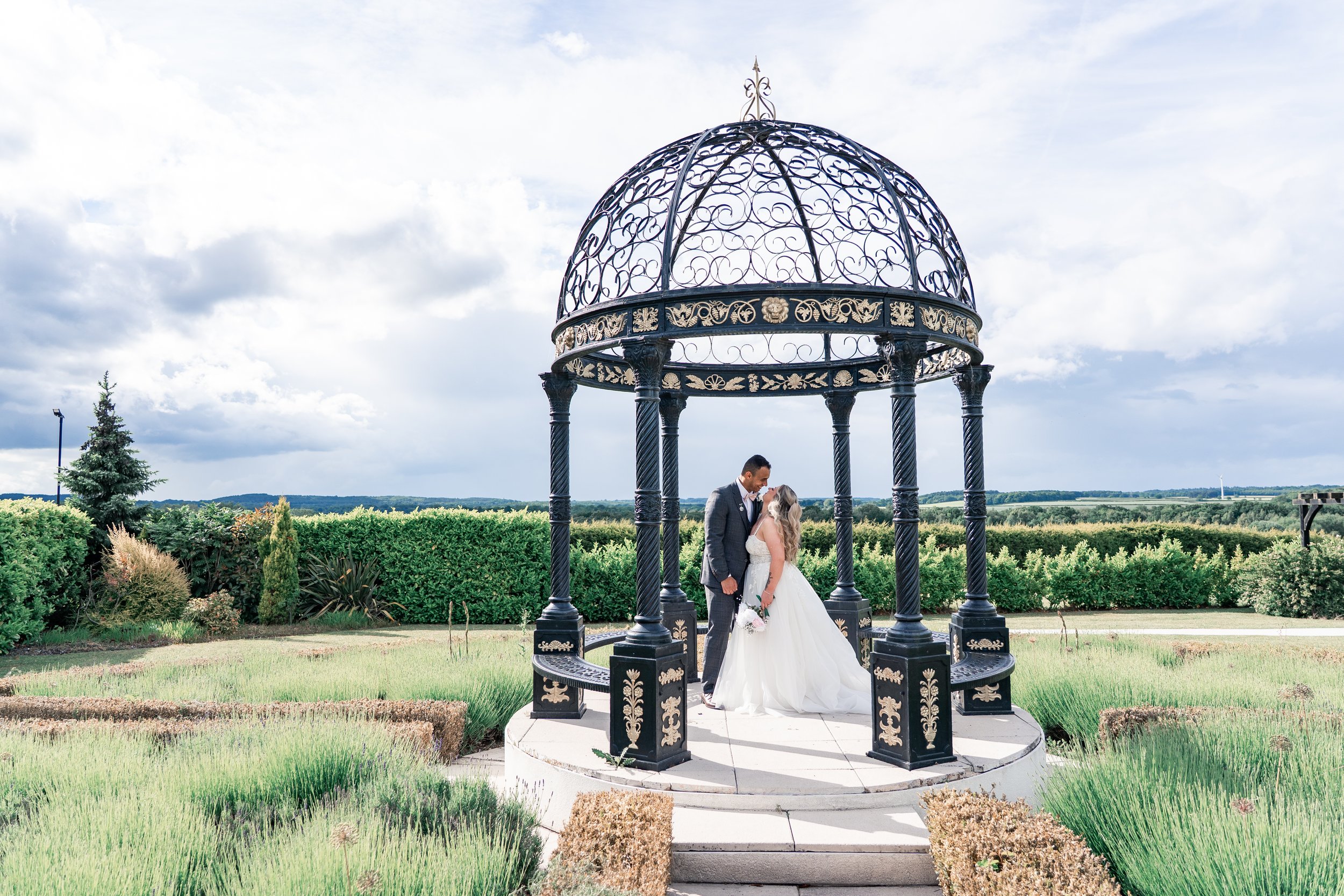 A bride and groom standing close together under a decorative black and gold gazebo outdoors on a cloudy day, with green bushes and trees in the background.