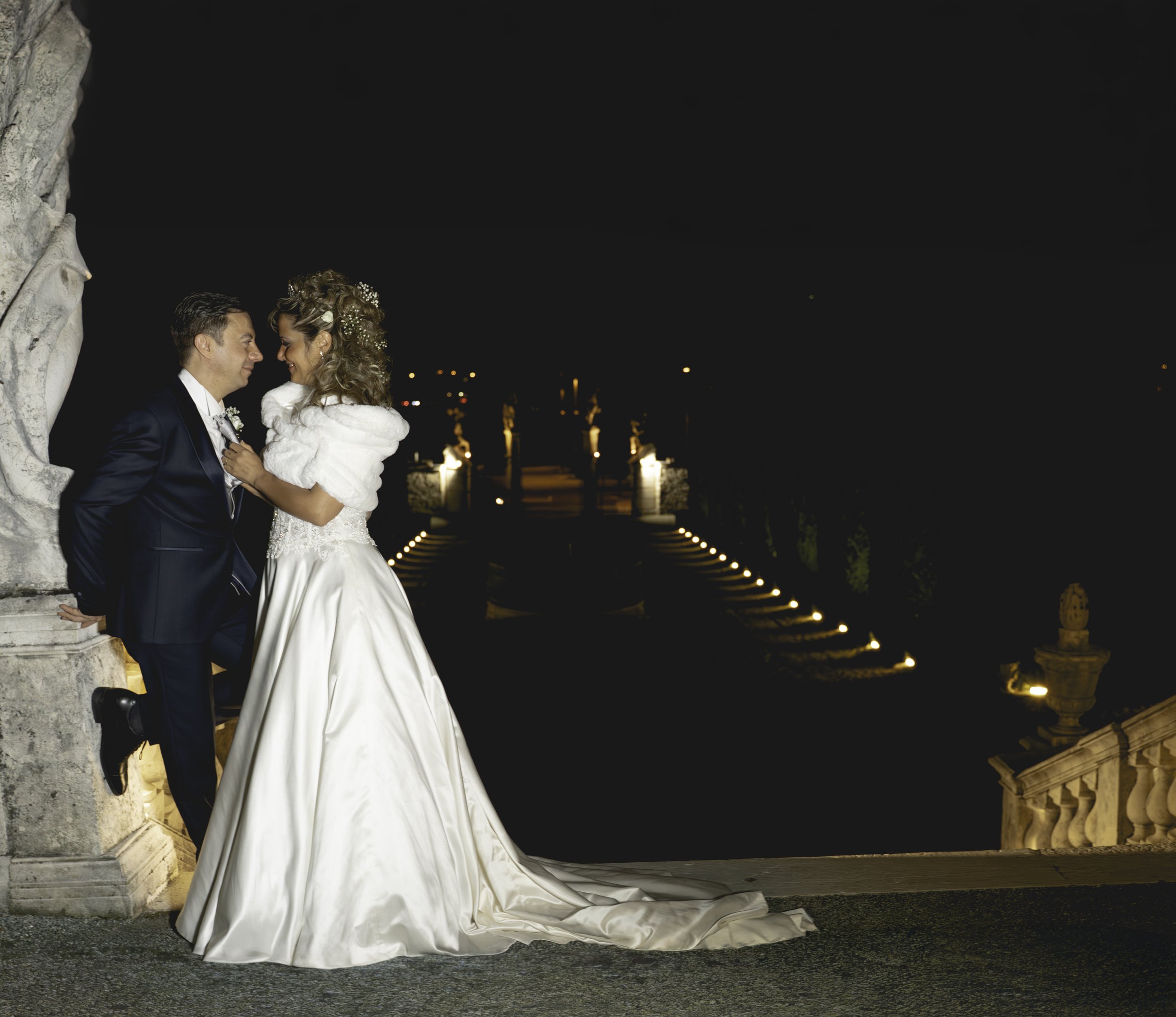 A newlywed couple at night, sharing a romantic moment, standing on a balcony with ornate stone balustrades and lit candles in the background.