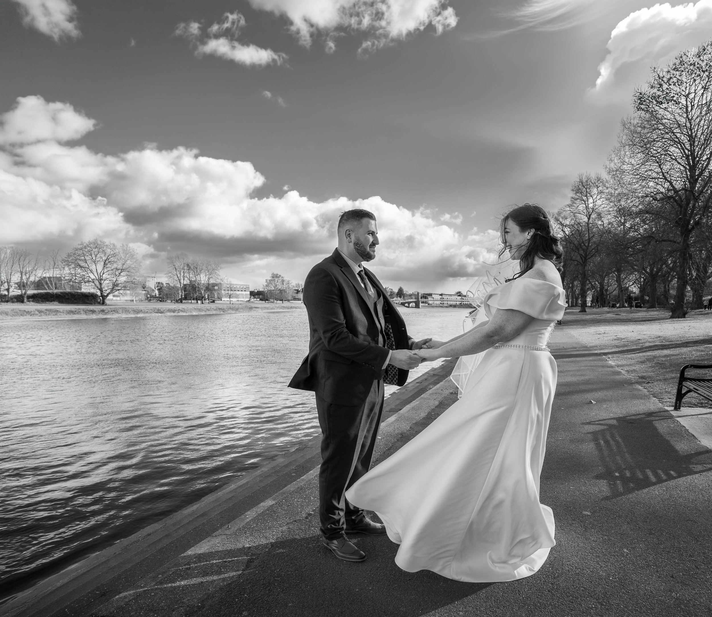 A black-and-white photo of a couple standing near a body of water outdoors on a cloudy day. The man is dressed in a suit and tie, facing a woman who is wearing a wedding gown. They are holding hands and looking at each other.