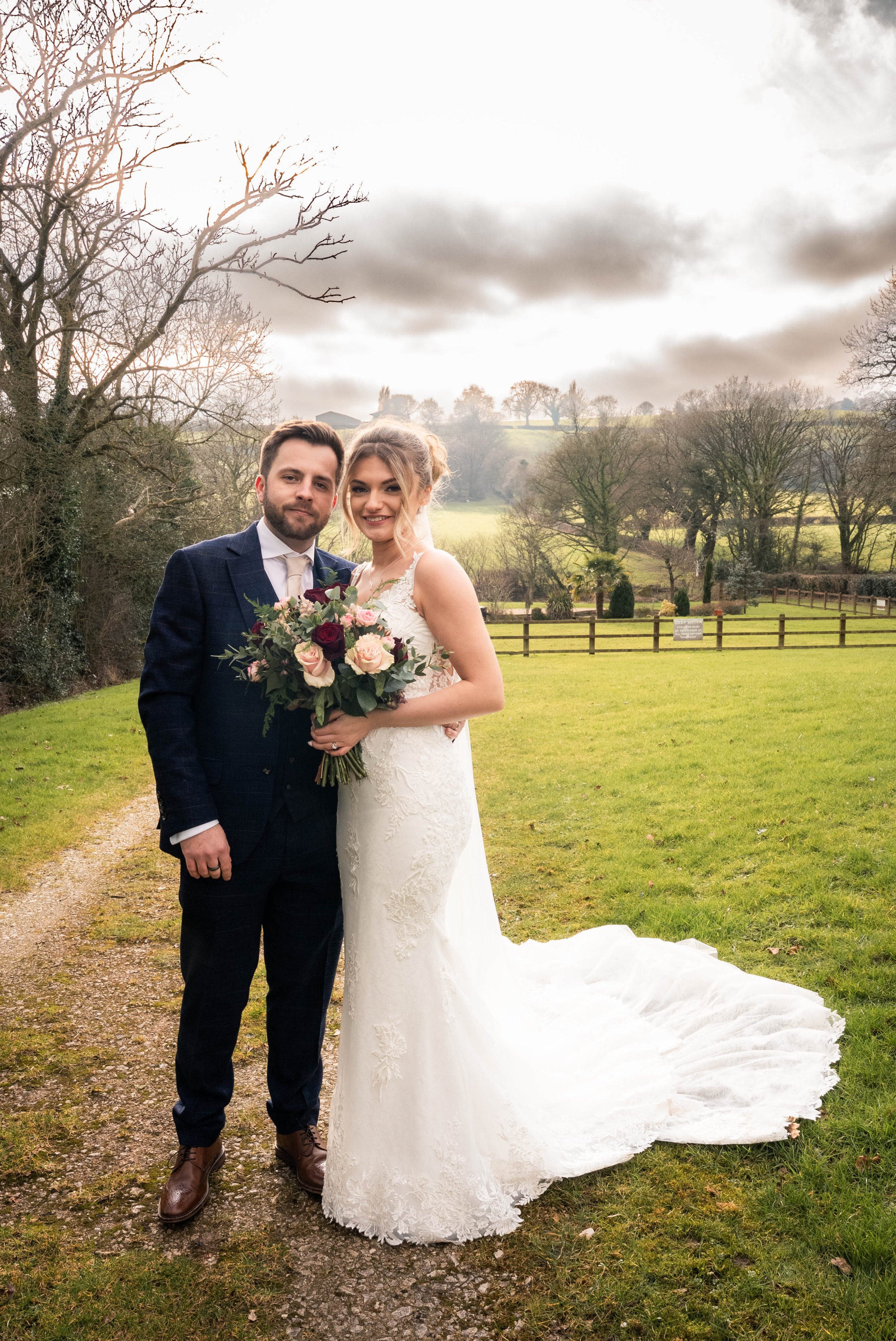 A newlywed couple standing outdoors on a grassy path, with the bride holding a bouquet of roses. The bride wears a white lace wedding gown with a train, and the groom wears a dark checkered suit with a white shirt and tie. They are smiling, with tree