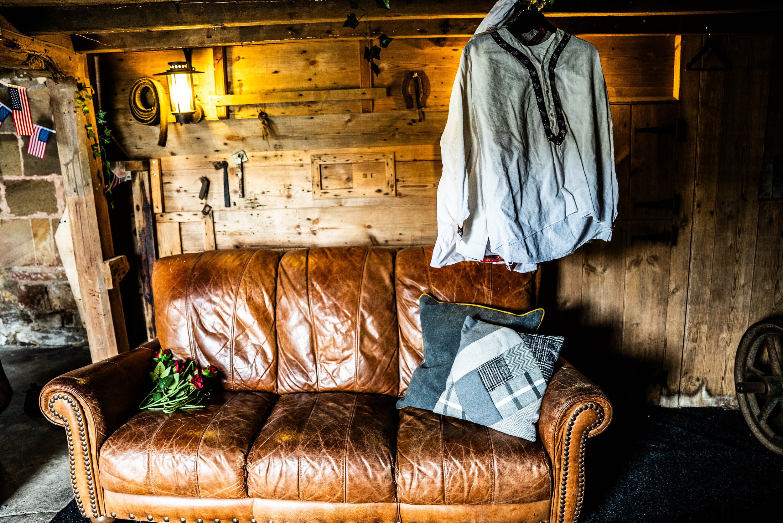 A brown leather couch with a cushion and a small bouquet of red flowers on it, within a rustic wooden room with hanging white shirt, wooden wall, and vintage decorations.