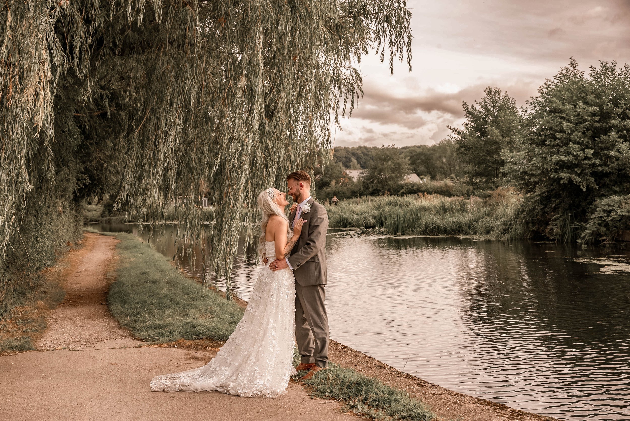 A bride and groom stand close together by a river at sunset, under a large tree with overhanging branches. The bride wears a white, lace wedding gown, and the groom wears a gray suit. They are smiling and gazing into each other's eyes.