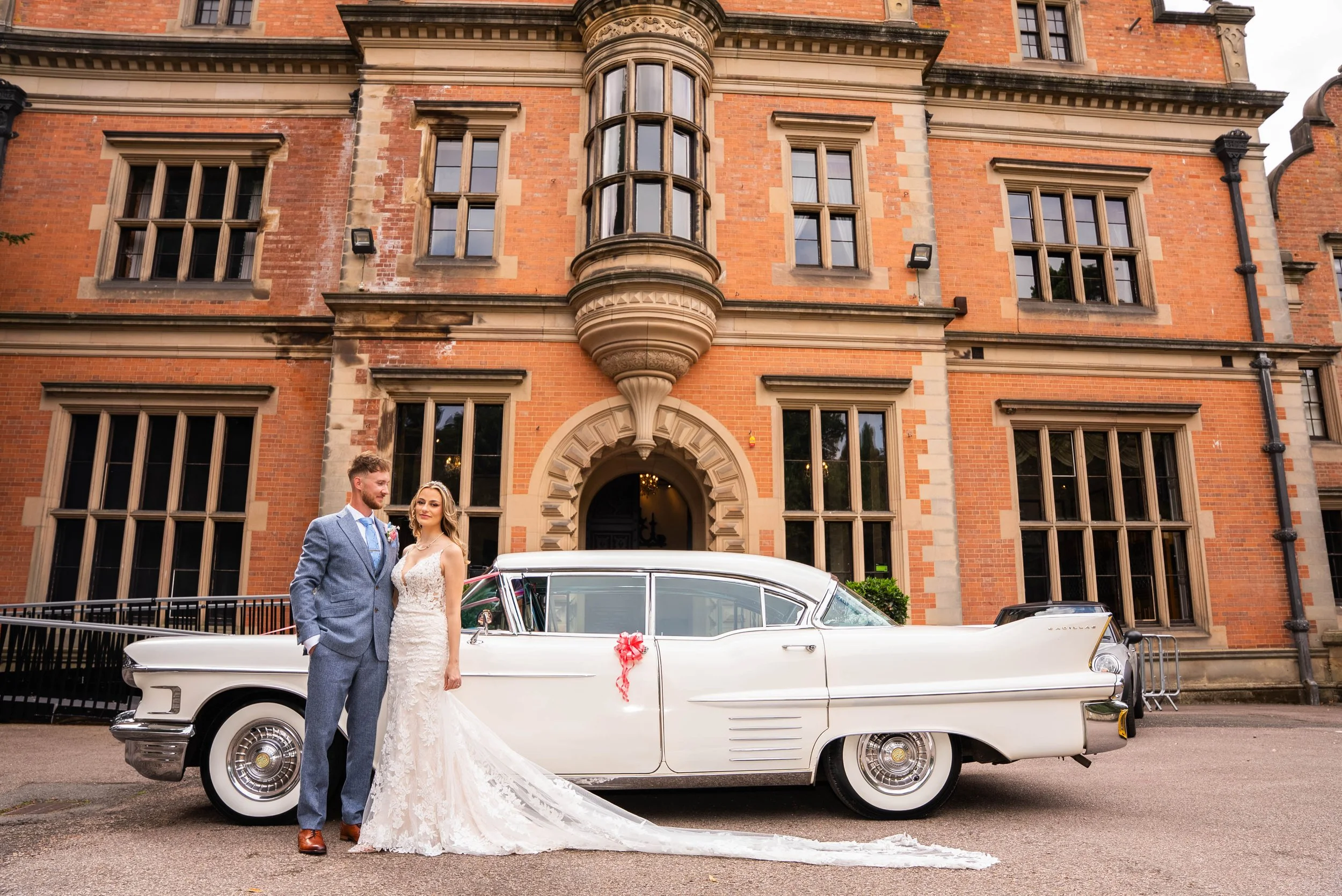 Newlywed couple in wedding attire standing in front of a vintage white Cadillac, decorated with a pink ribbon, outside a large brick historic building.