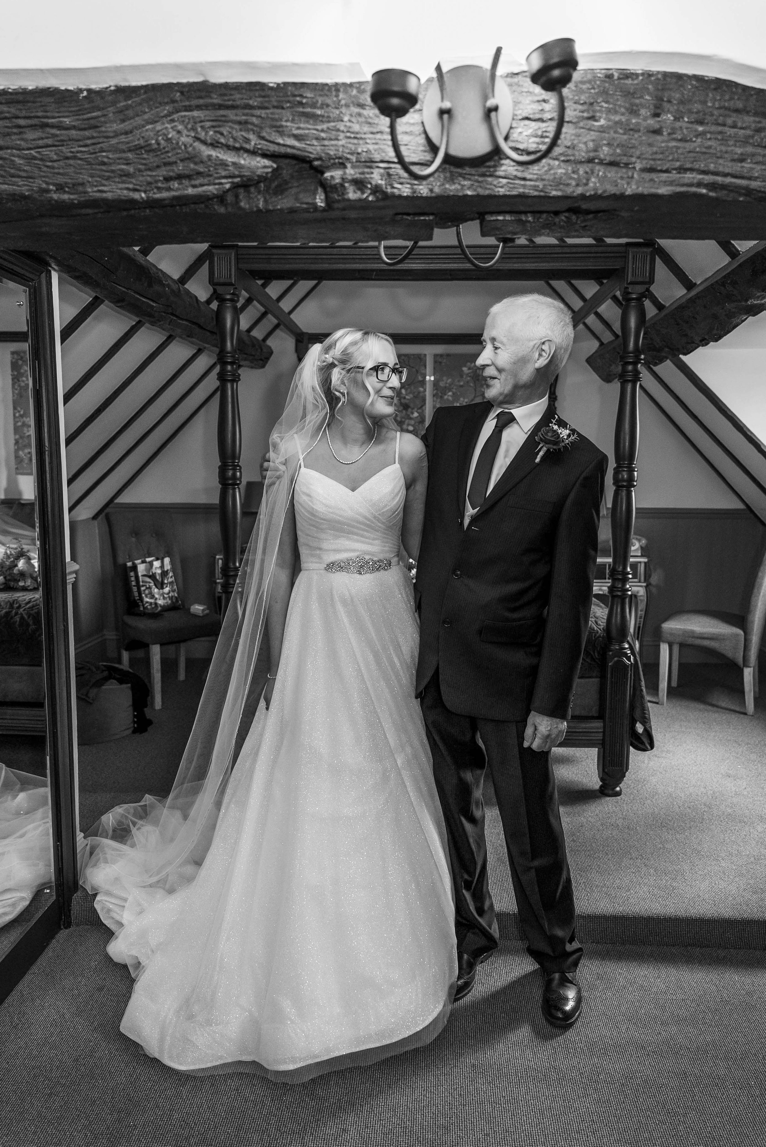 A bride with glasses and earrings smiling at an elderly man in a suit, in an indoor setting with wooden beams and furniture, during a wedding event.
