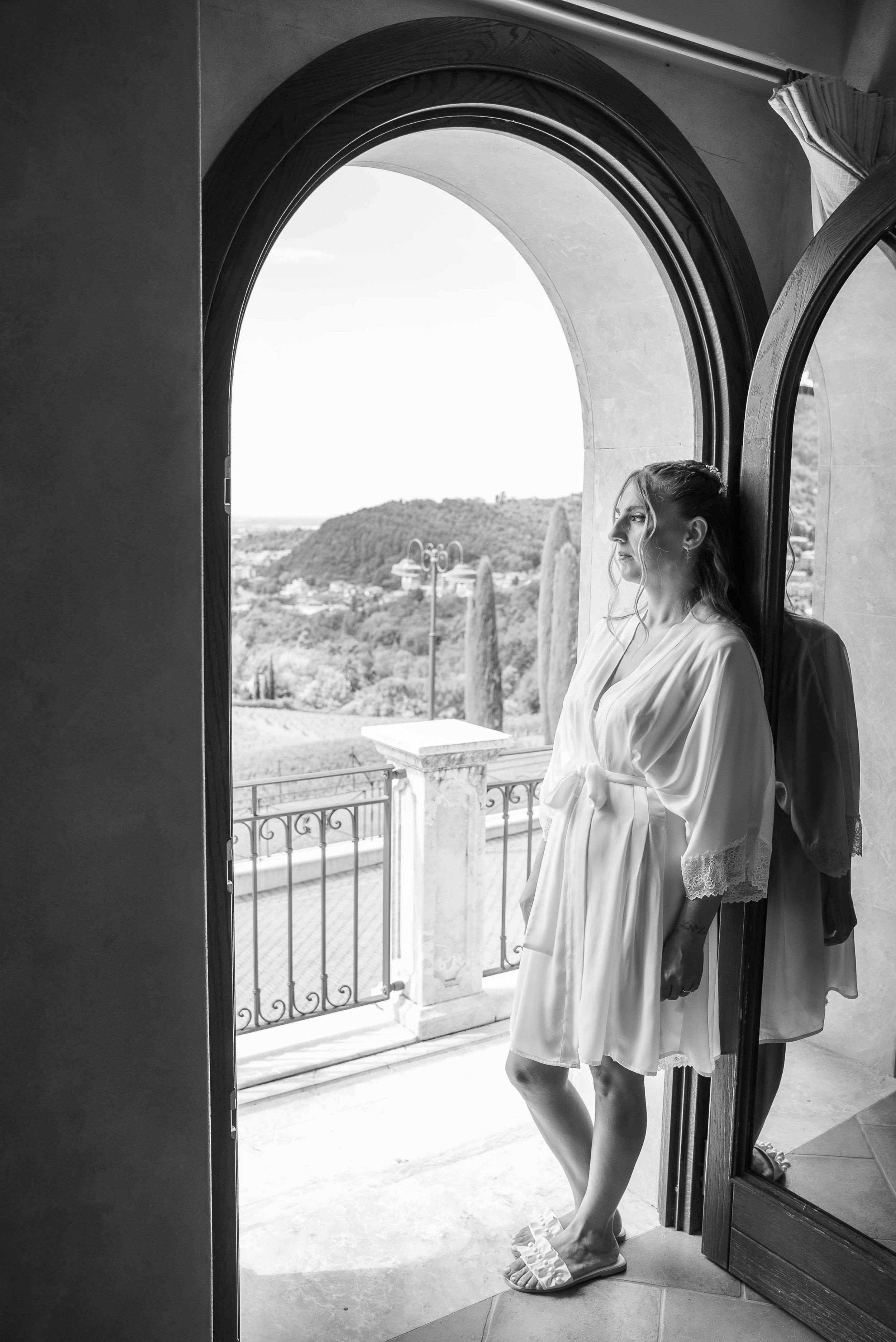 A woman in a white robe and slippers stands at an open door, holding a person's hand. She is looking outside at a view of trees, hills, and a cloudy sky.