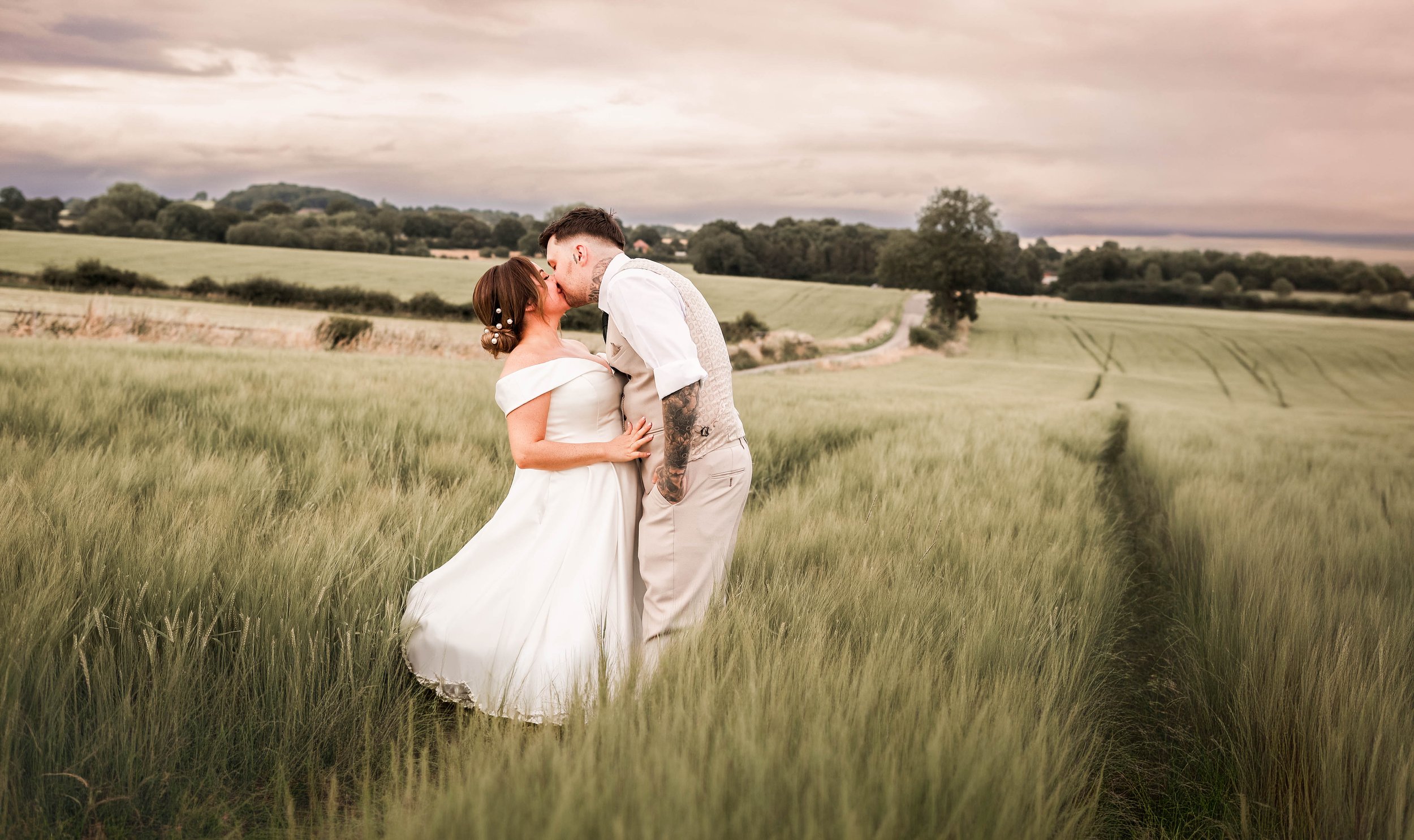 A bride and groom share a kiss in a green field with tall grass, under a cloudy sky, in a rural landscape.