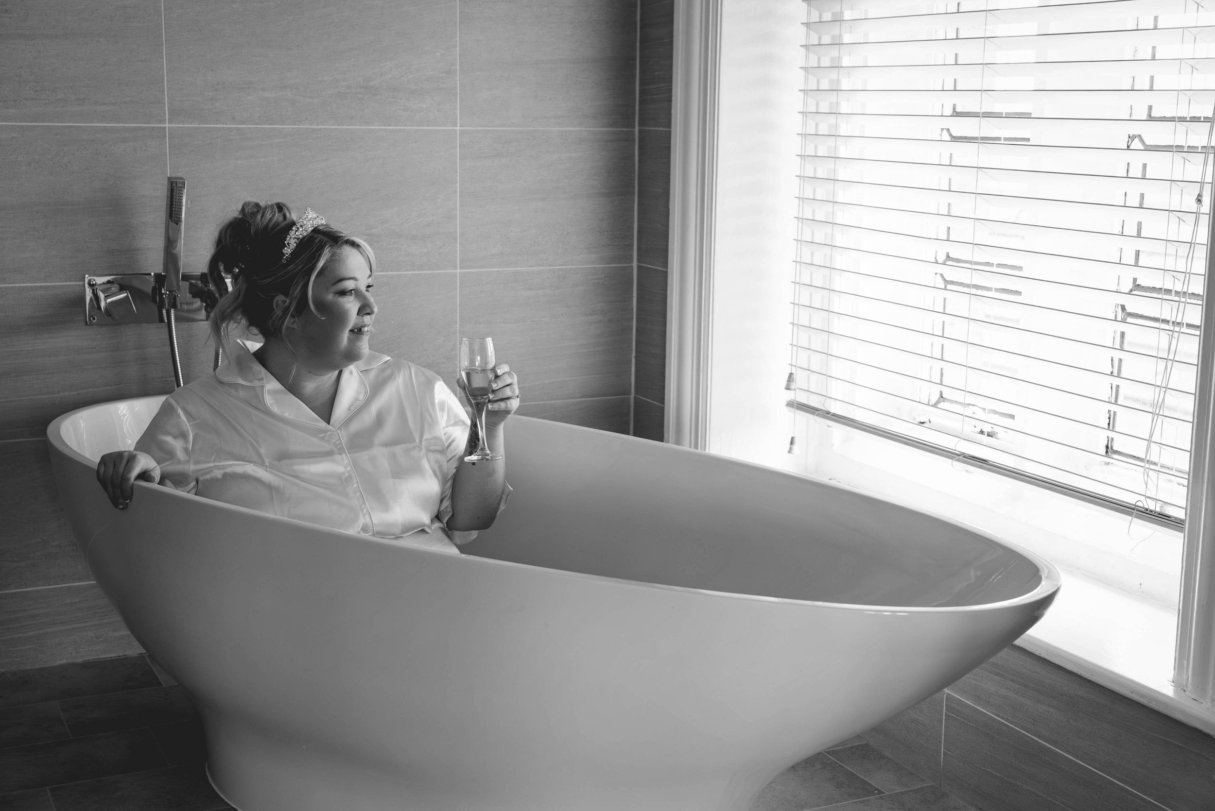 A woman wearing a tiara and a satin robe relaxes in a clawfoot bathtub, holding a glass of champagne, in a modern bathroom with large window blinds.