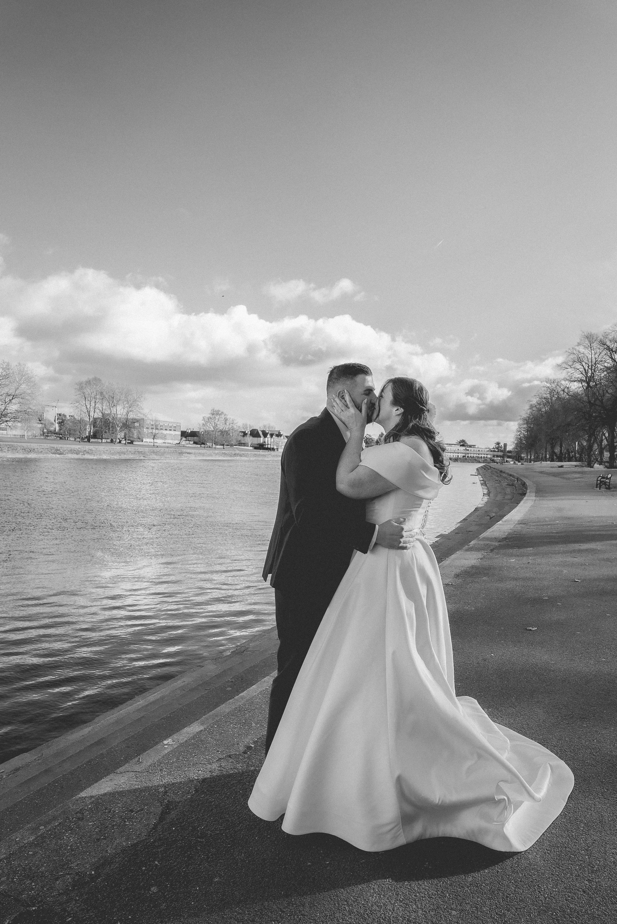 A black and white photo of a wedding couple kissing by a river, with trees and buildings in the background.